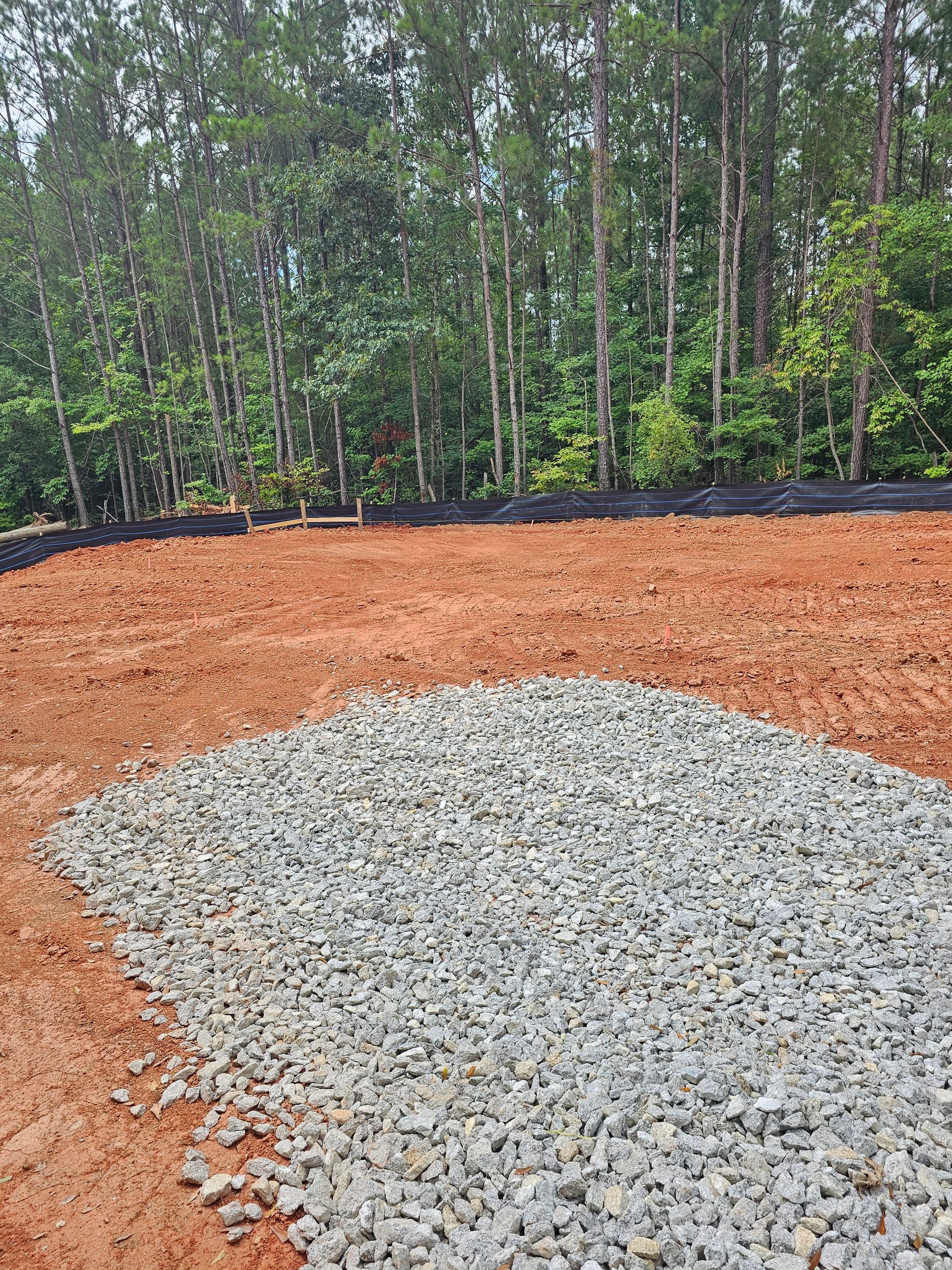 Cleared red soil with a pile of gray gravel in front of a treeline.