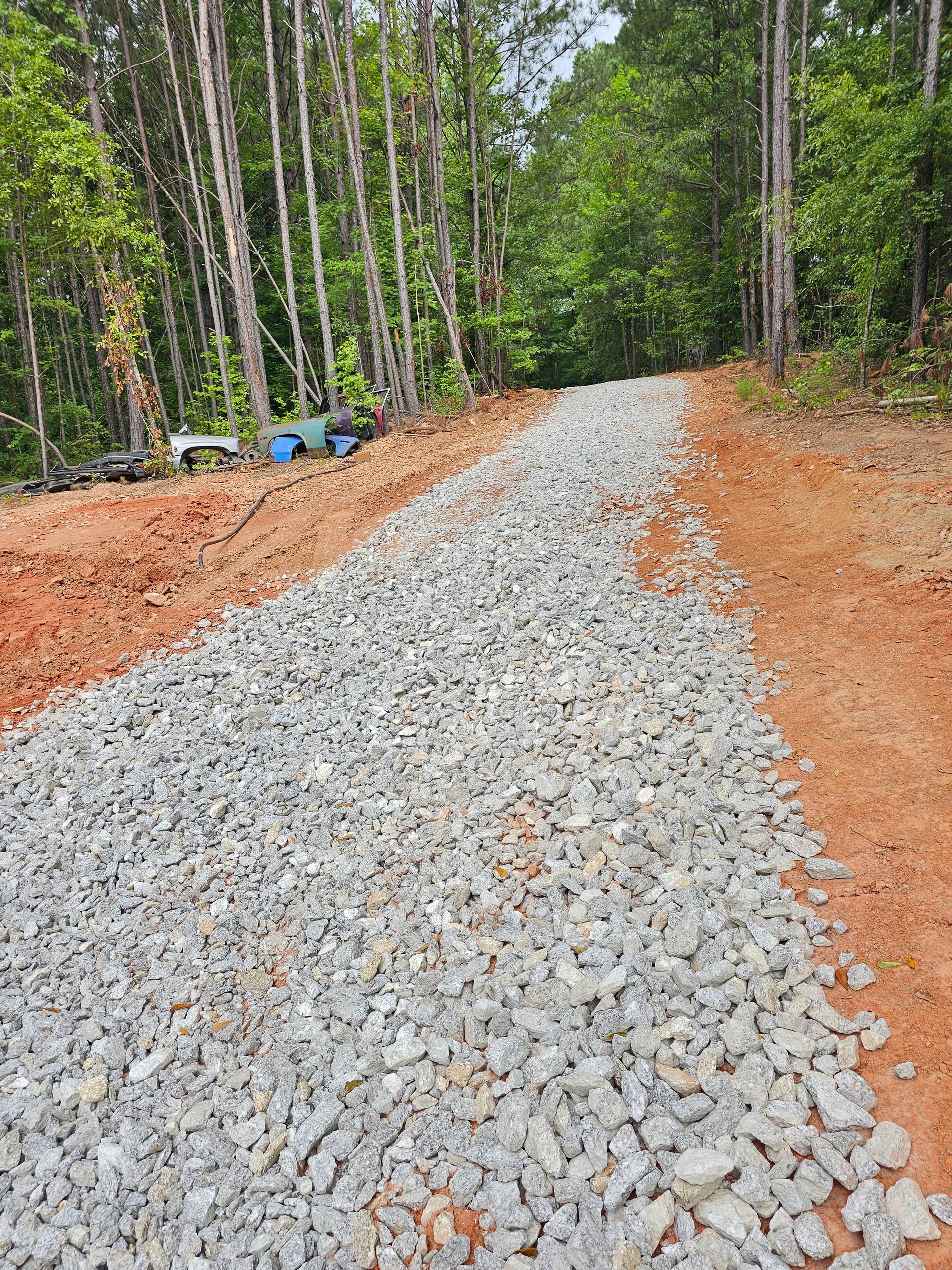 Gravel road through a forest. Red dirt edges the path of gray rock. Green trees line the sides of the road.