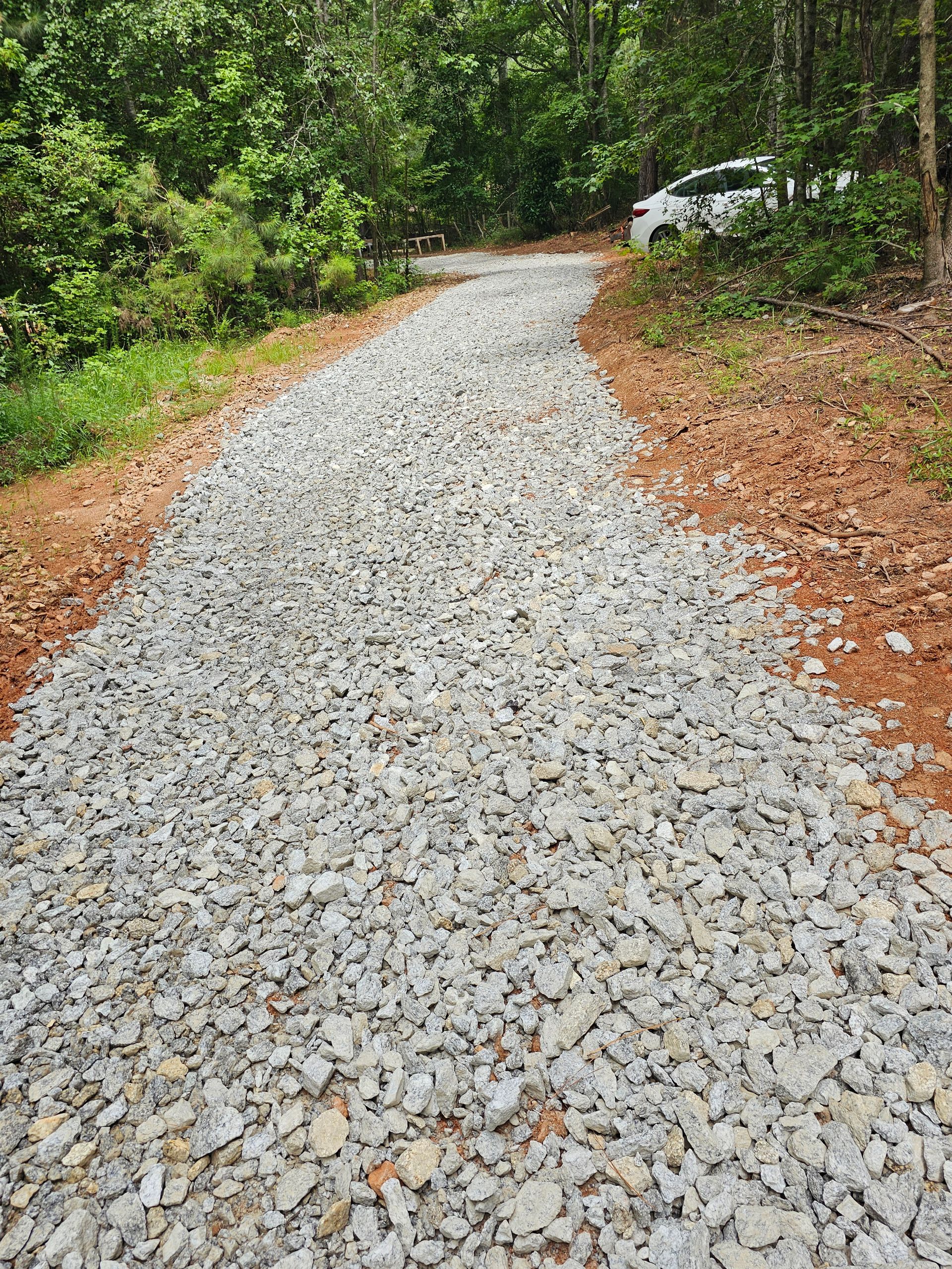A gravel driveway curves uphill through a wooded area; a white car is parked at the top.
