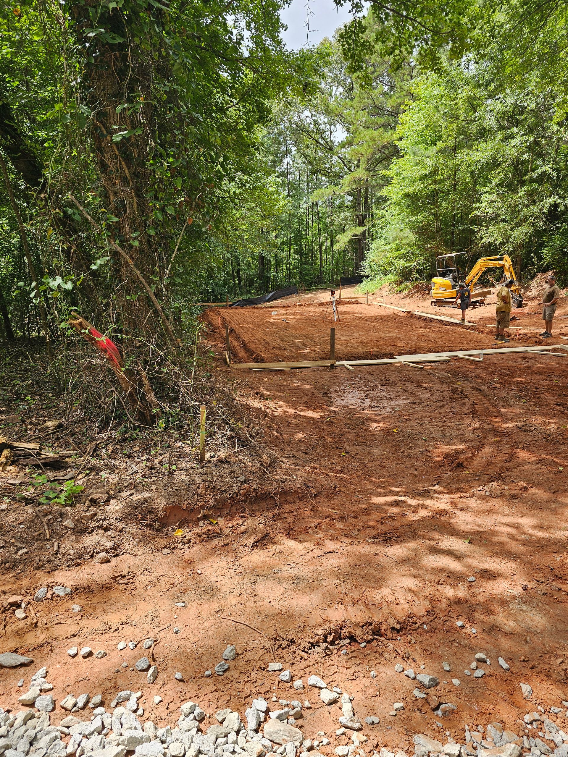 Cleared construction site with dirt and gravel, surrounded by trees. An excavator and workers are present.