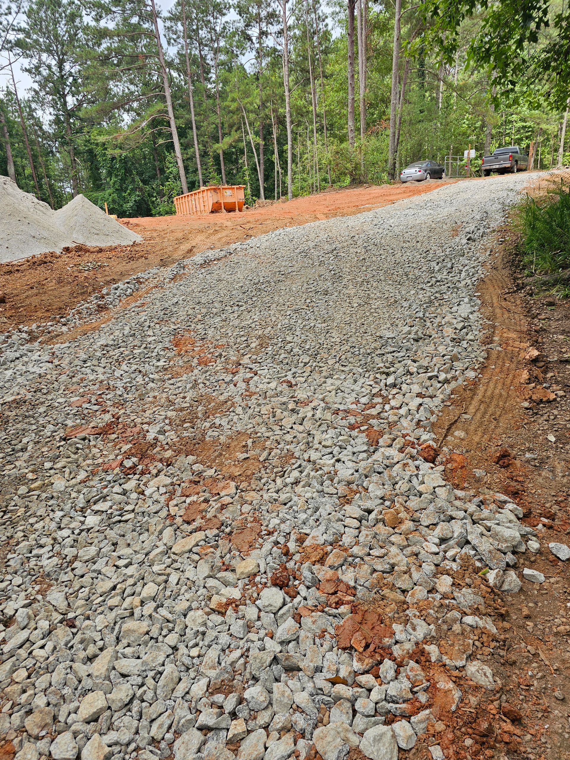 Gravel road under construction, with red soil and pile of rocks. In background, trees and building materials.