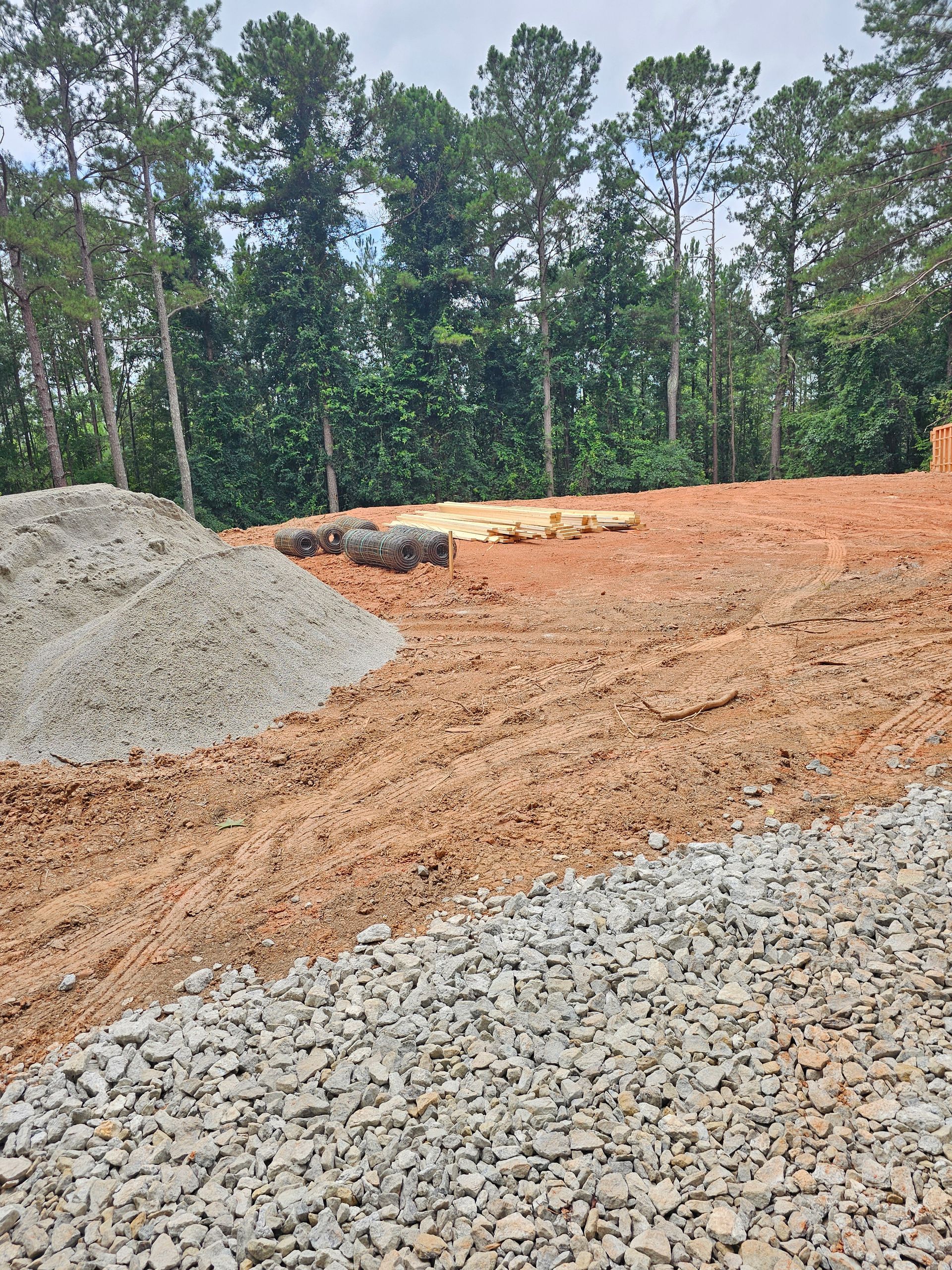 Construction site with piles of gravel and dirt in a clearing surrounded by trees.