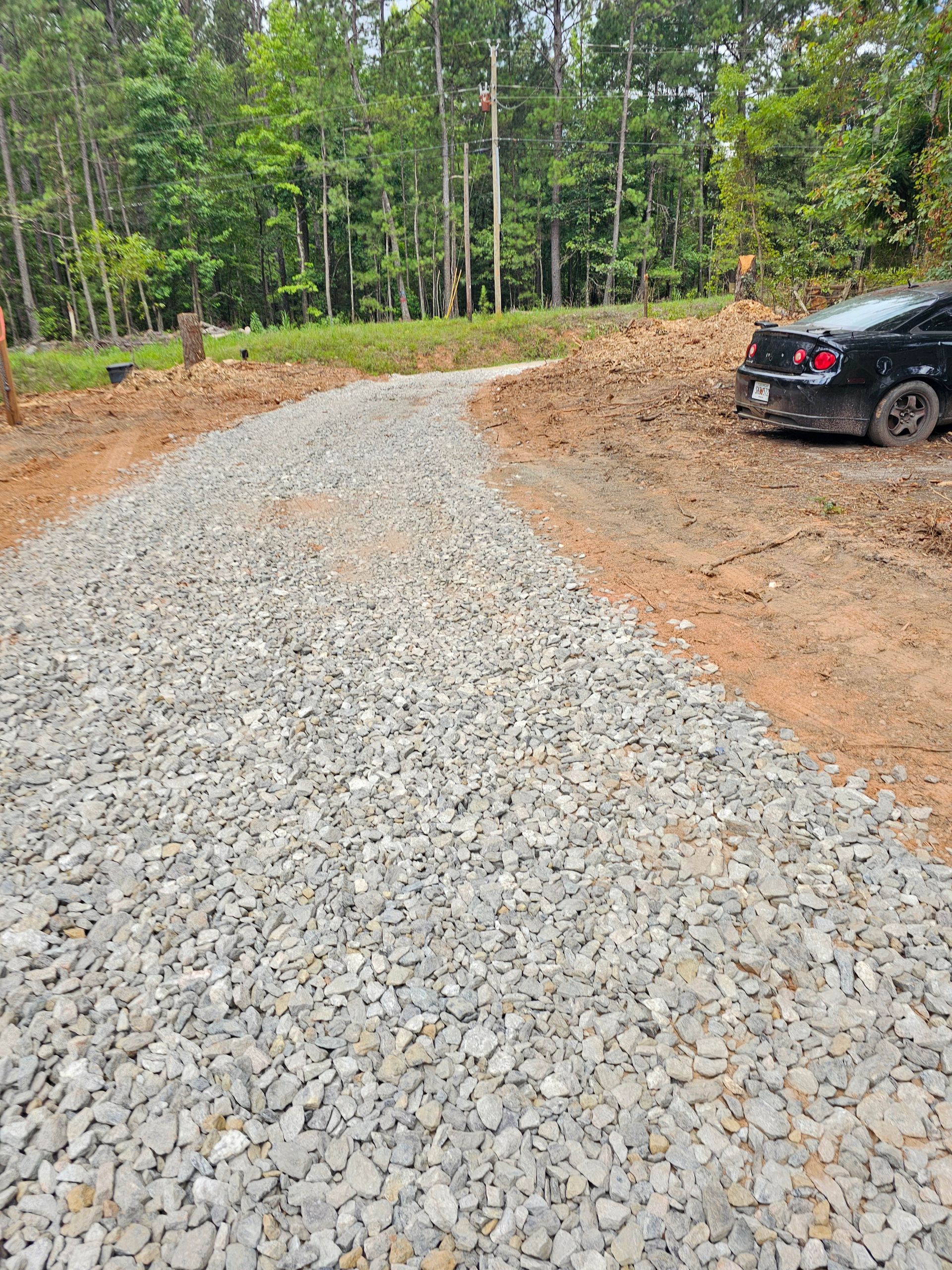 Gravel driveway curving through an undeveloped lot. A black car is parked to the side. Trees and forest in the background.