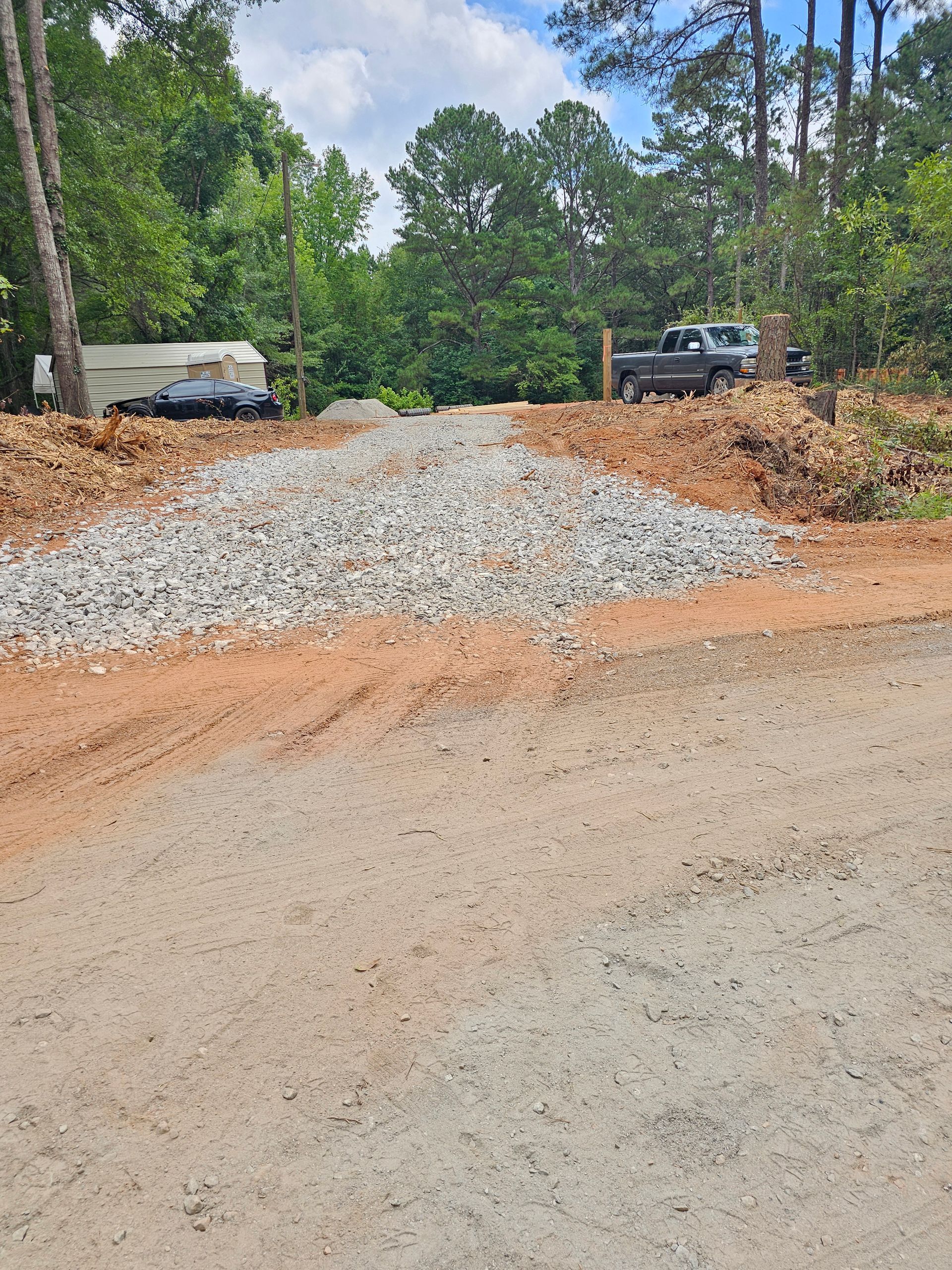 Dirt road entrance to property with gravel and truck visible. Trees and cloudy sky in background.