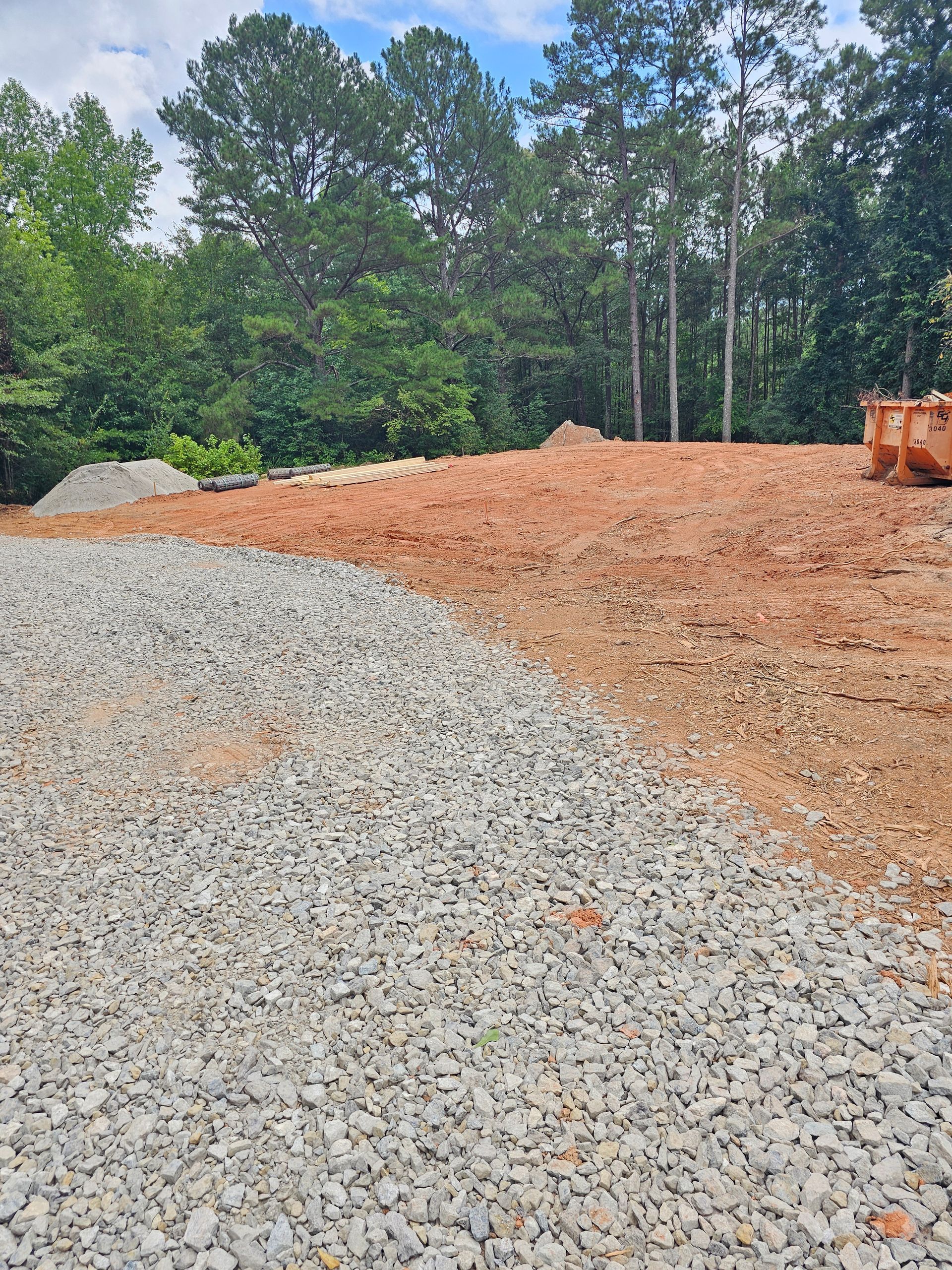 Gravel driveway transitions to a dirt lot. Trees in background, orange bin at right.