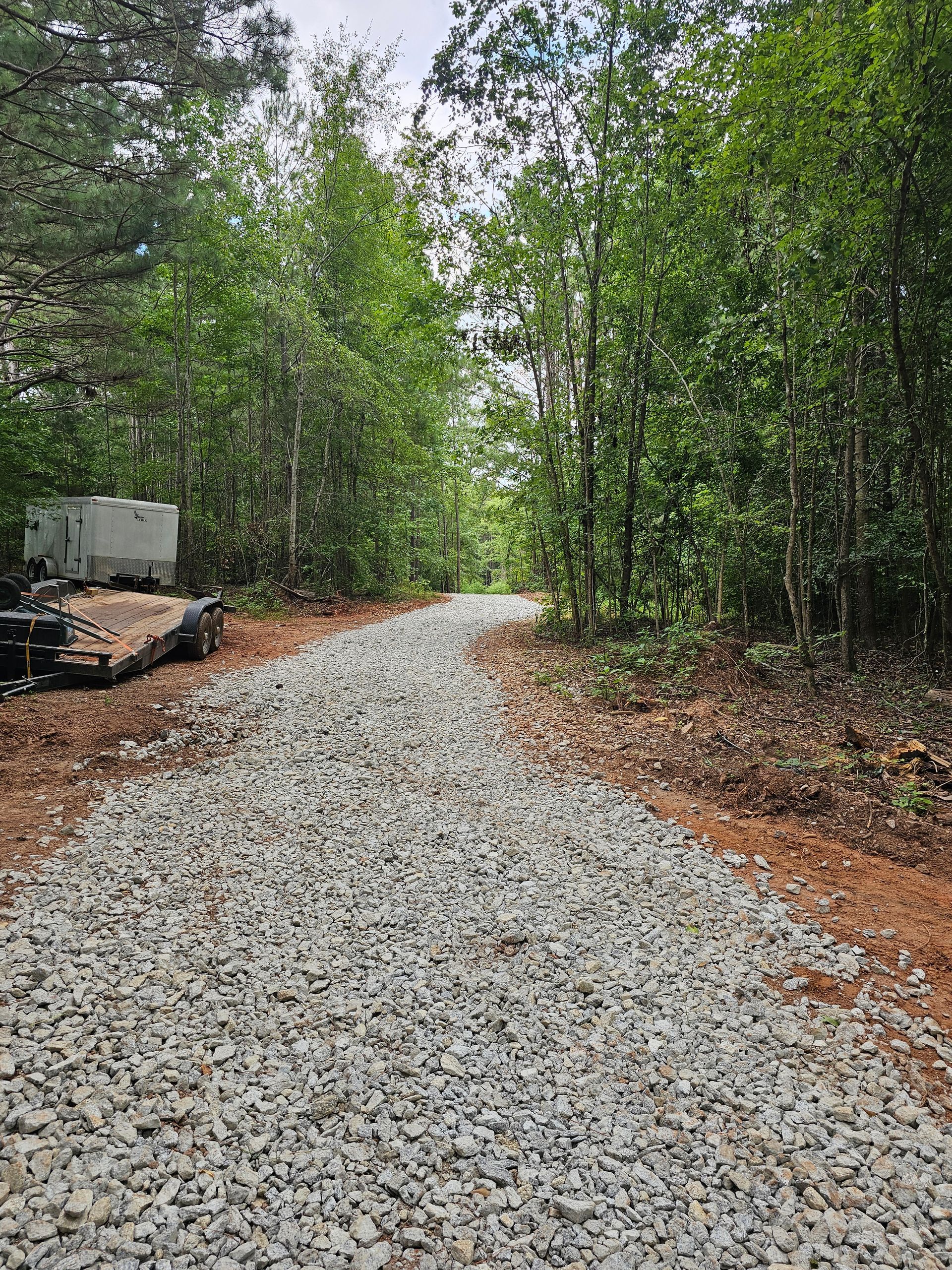 Gravel path through a forest, trees on either side. A concrete structure is visible on the left.