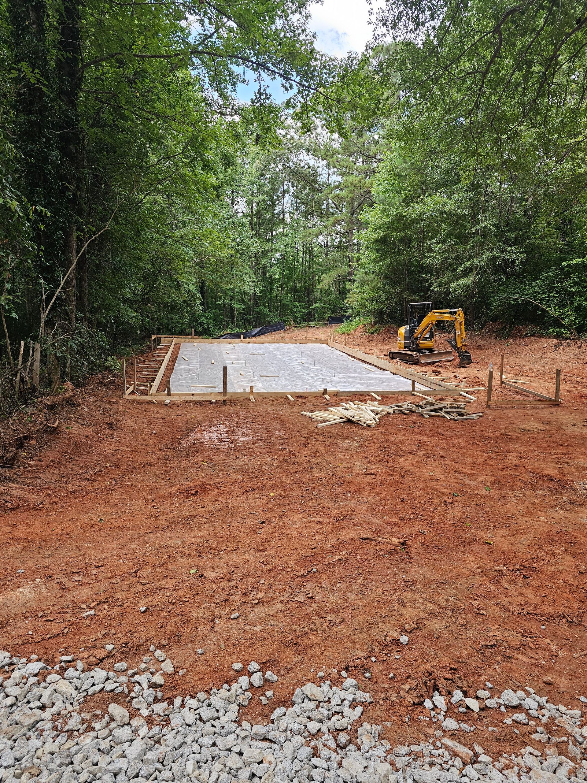 Construction site with a concrete foundation, excavator, and surrounding trees.