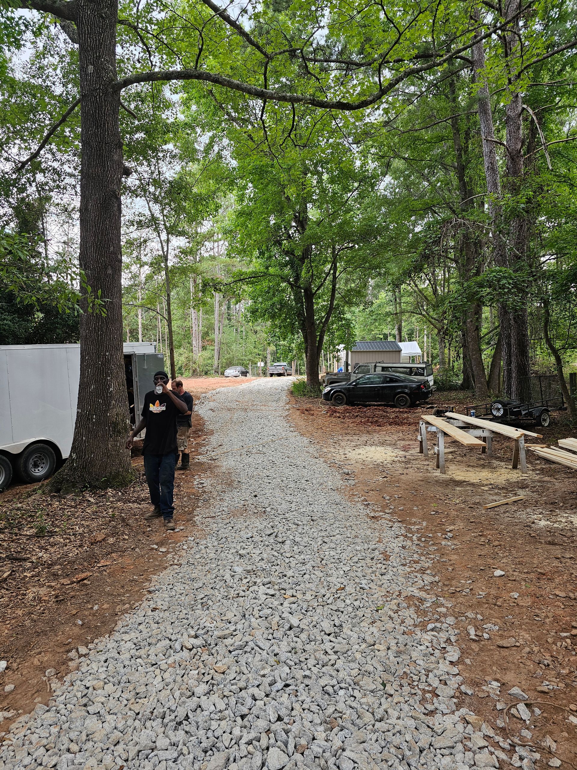 Gravel path through a wooded area. Two people walk towards camera. Trailers and vehicles visible.