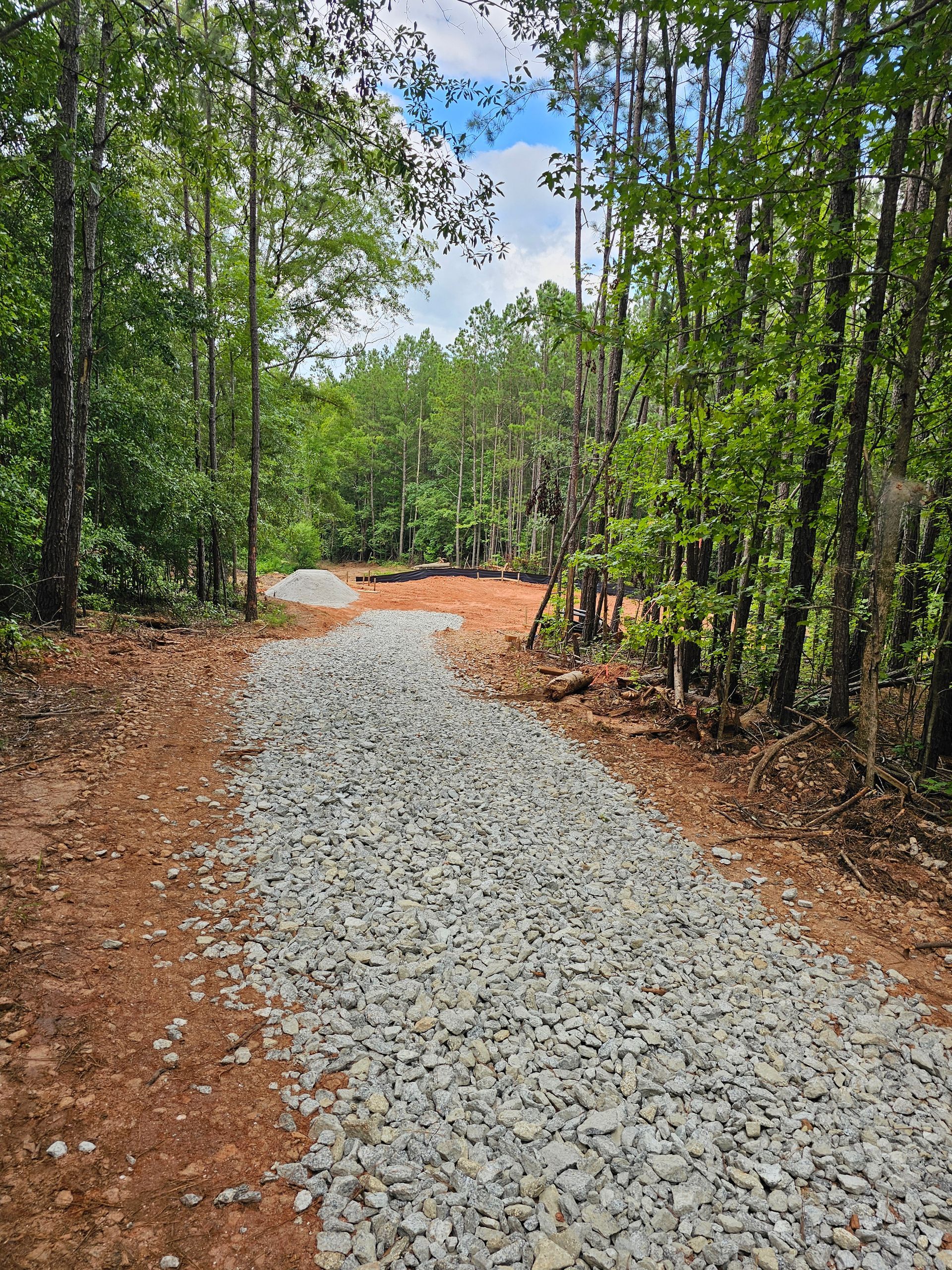 Gravel path through a wooded area; trees line the sides, leading to a cleared area under a partly cloudy sky.
