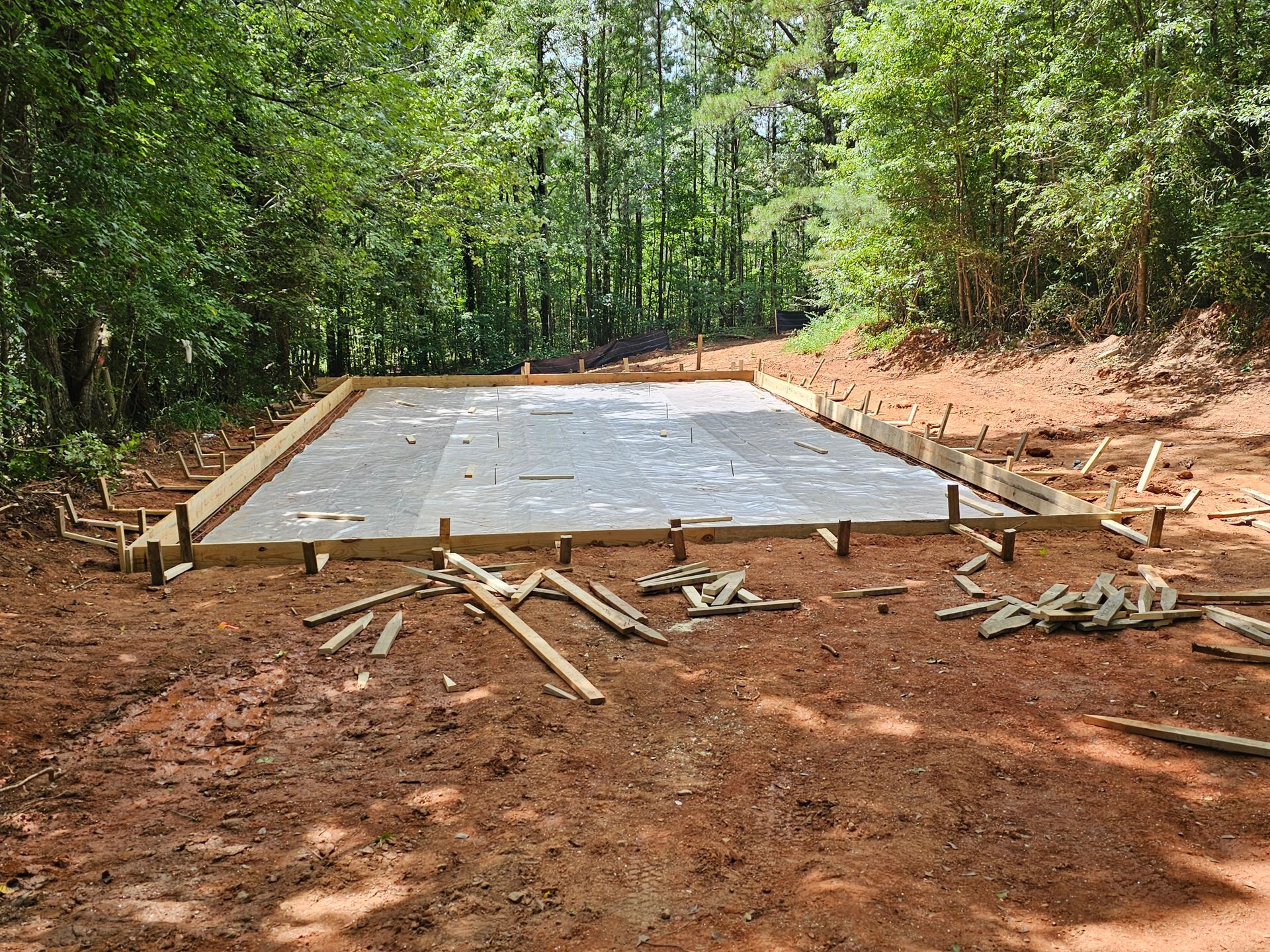 Freshly poured concrete slab with wooden framing on dirt ground in a wooded area.