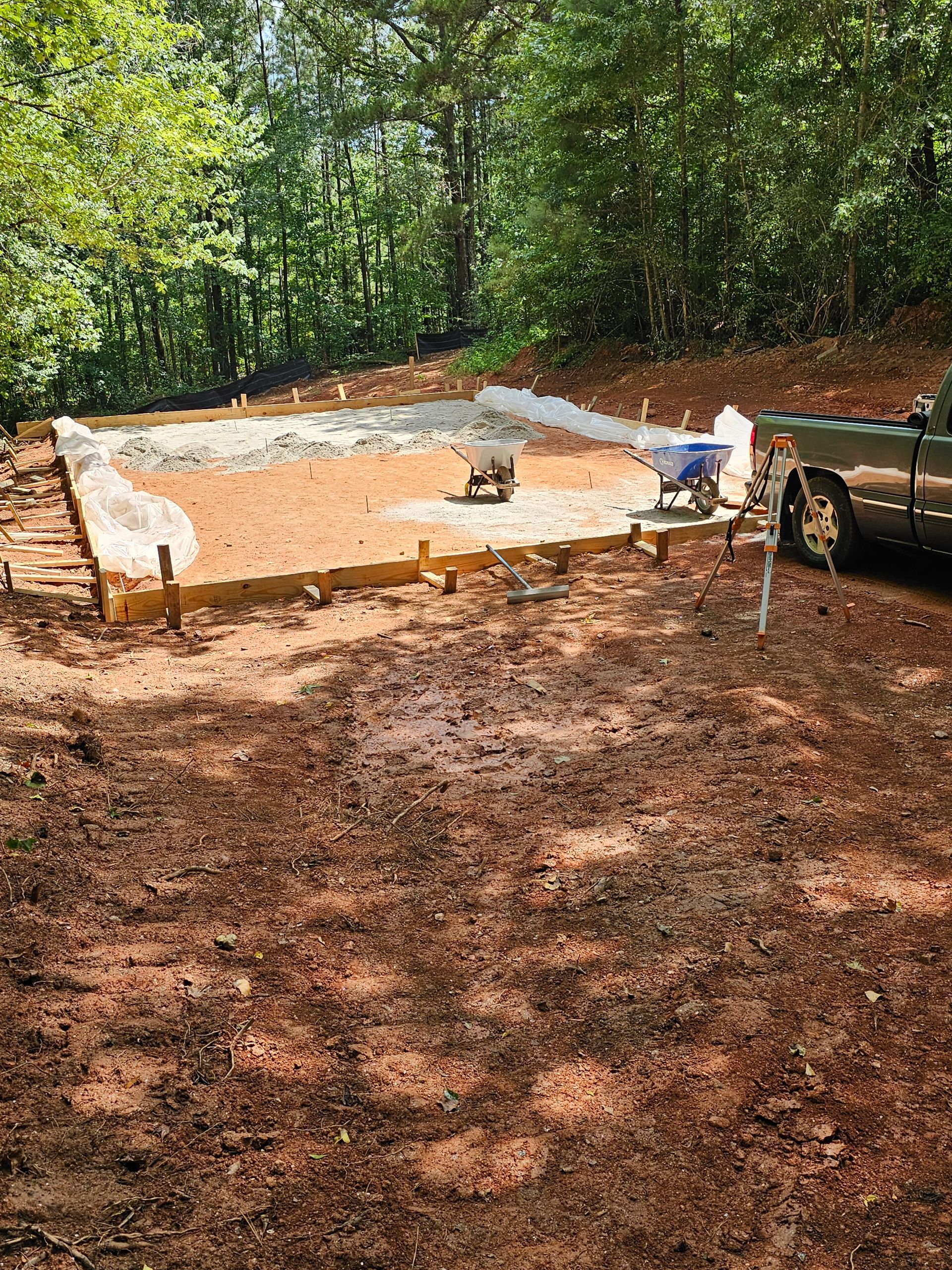 Construction site: Foundation outline with gravel, surrounded by dirt and forest, a truck and wheelbarrow present.