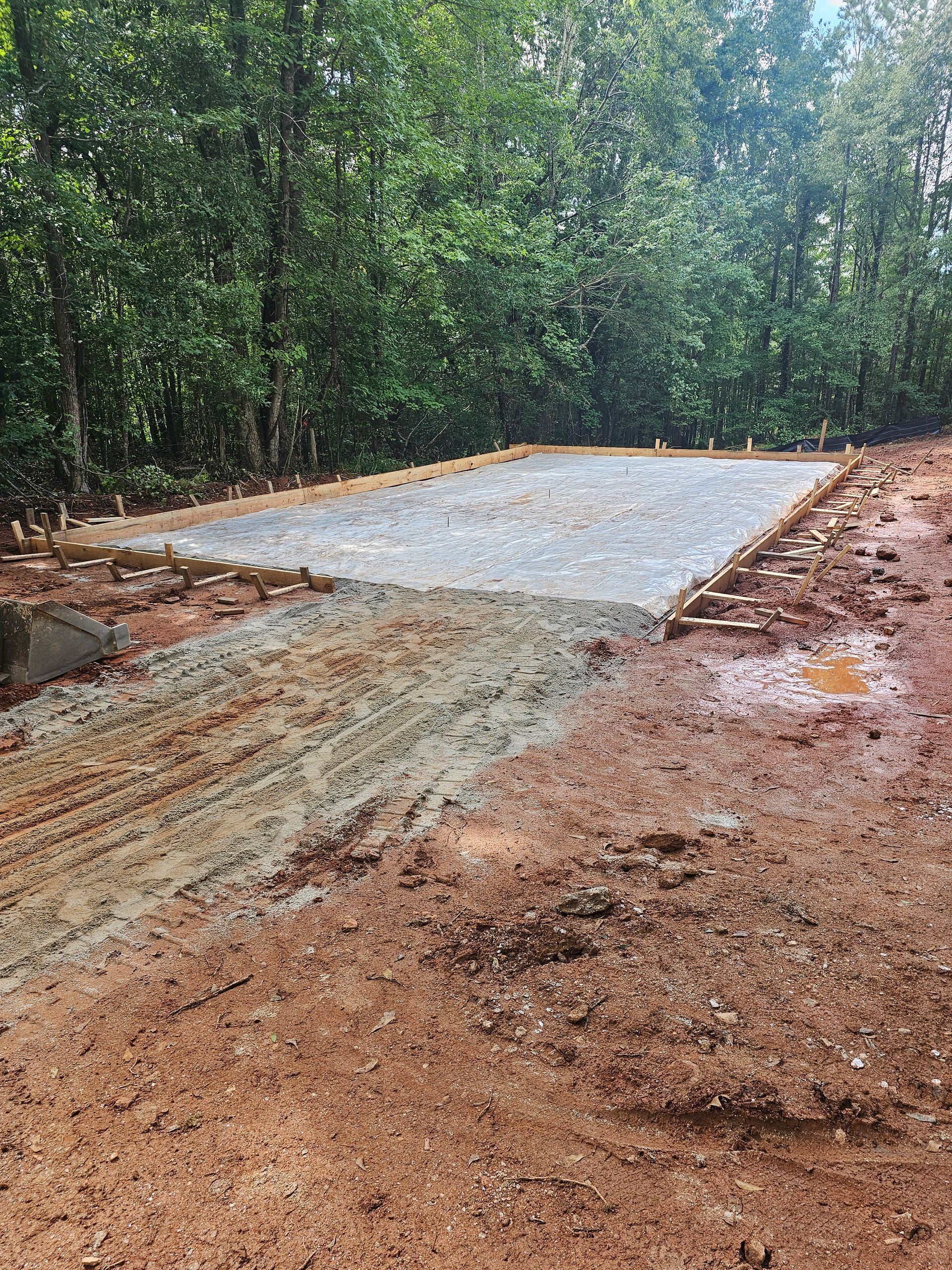 Construction site: gravel foundation bordered by wood, muddy foreground, surrounded by trees.