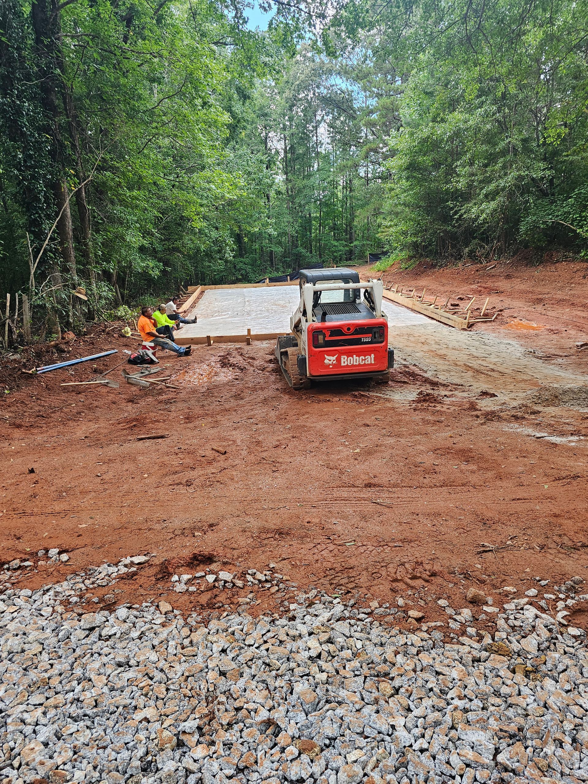 Construction site with red Bobcat skid-steer, concrete pad, and worker in orange vest, surrounded by trees.