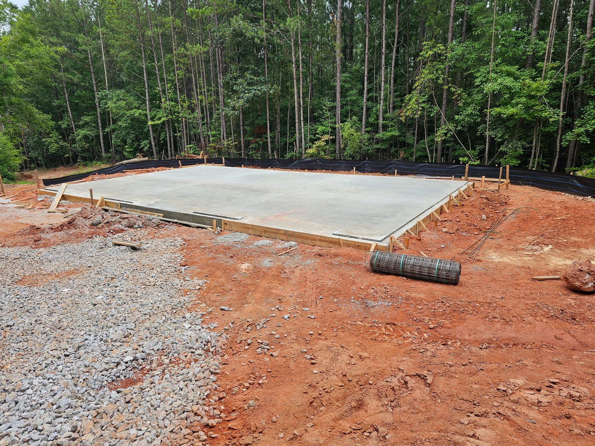 A concrete slab foundation with wooden framing on red clay ground, with a forest in the background.