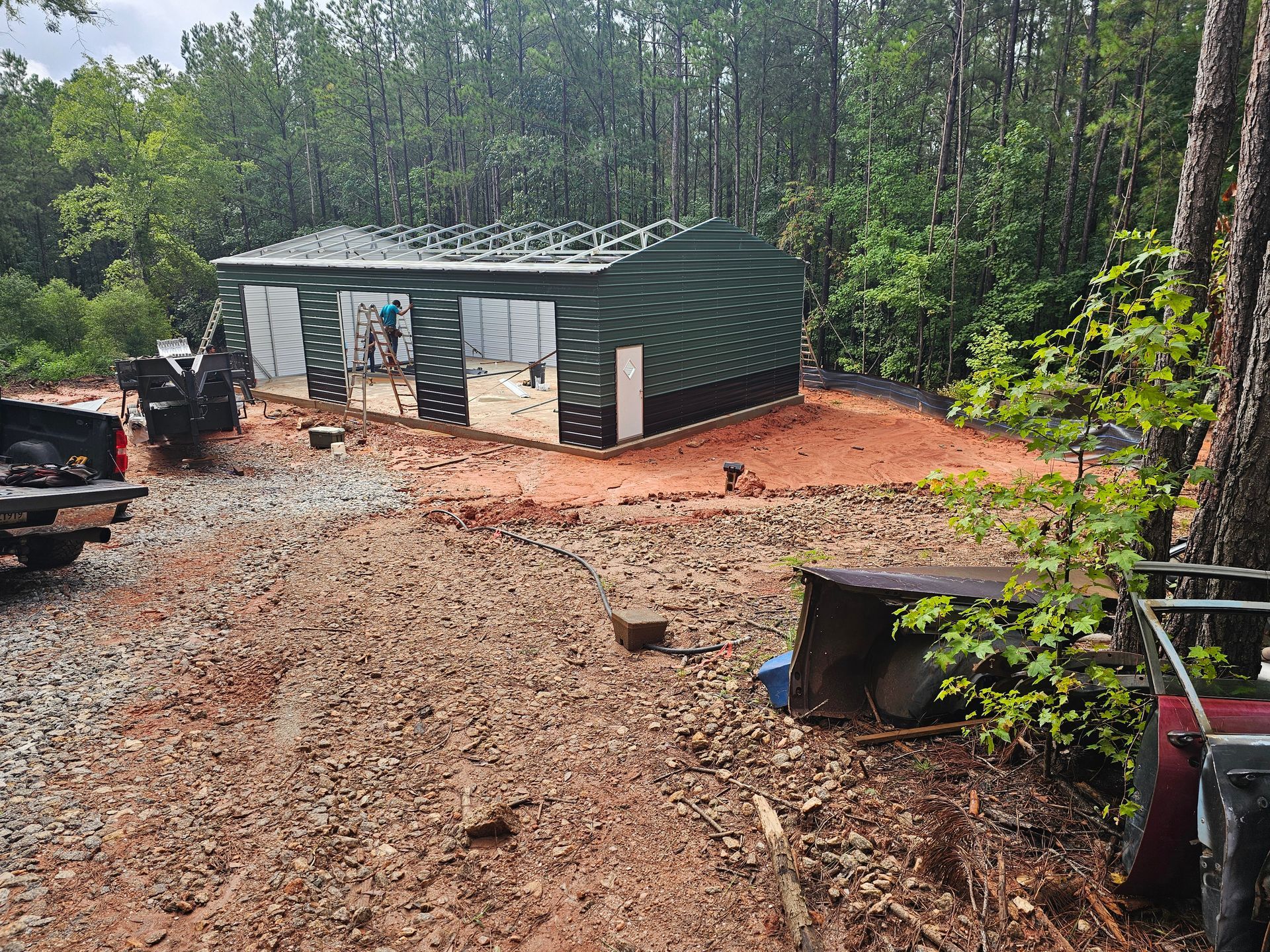 Green metal building under construction in a wooded area; dirt and gravel ground.