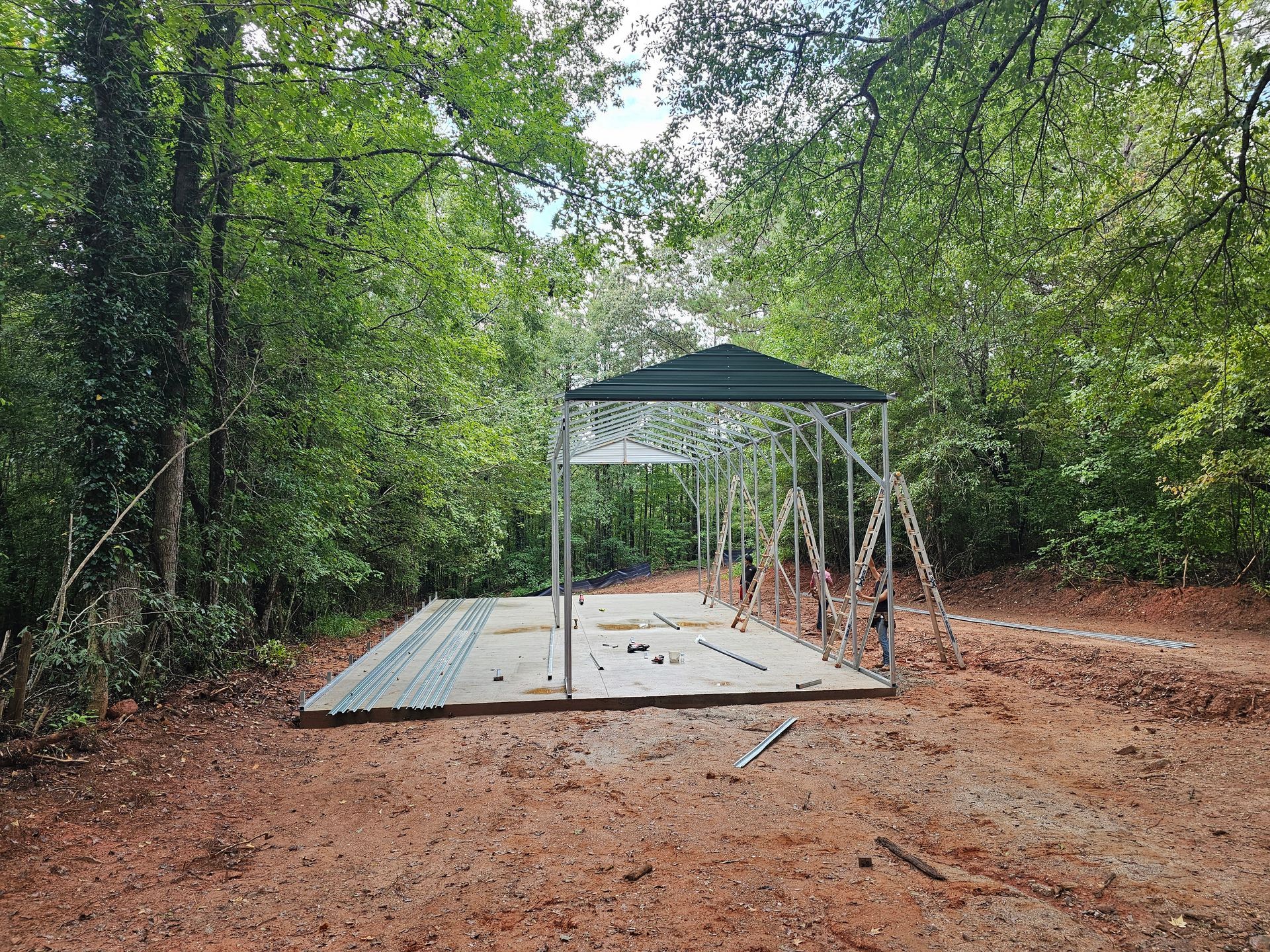 A partially constructed metal shelter with a green roof, set in a wooded area.