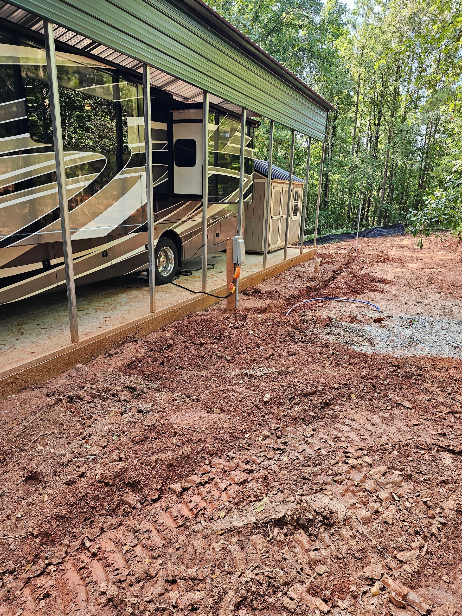 An RV parked under a green metal carport. Dirt and gravel driveway with a treed background.