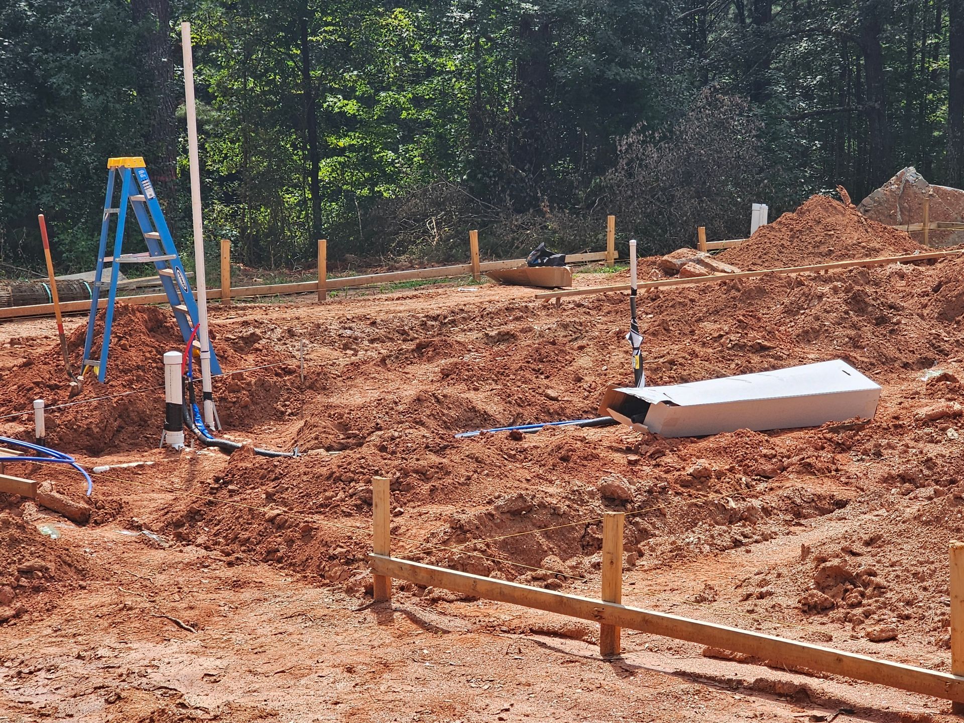 Construction site with graded red dirt, wooden stakes, and blue ladder.