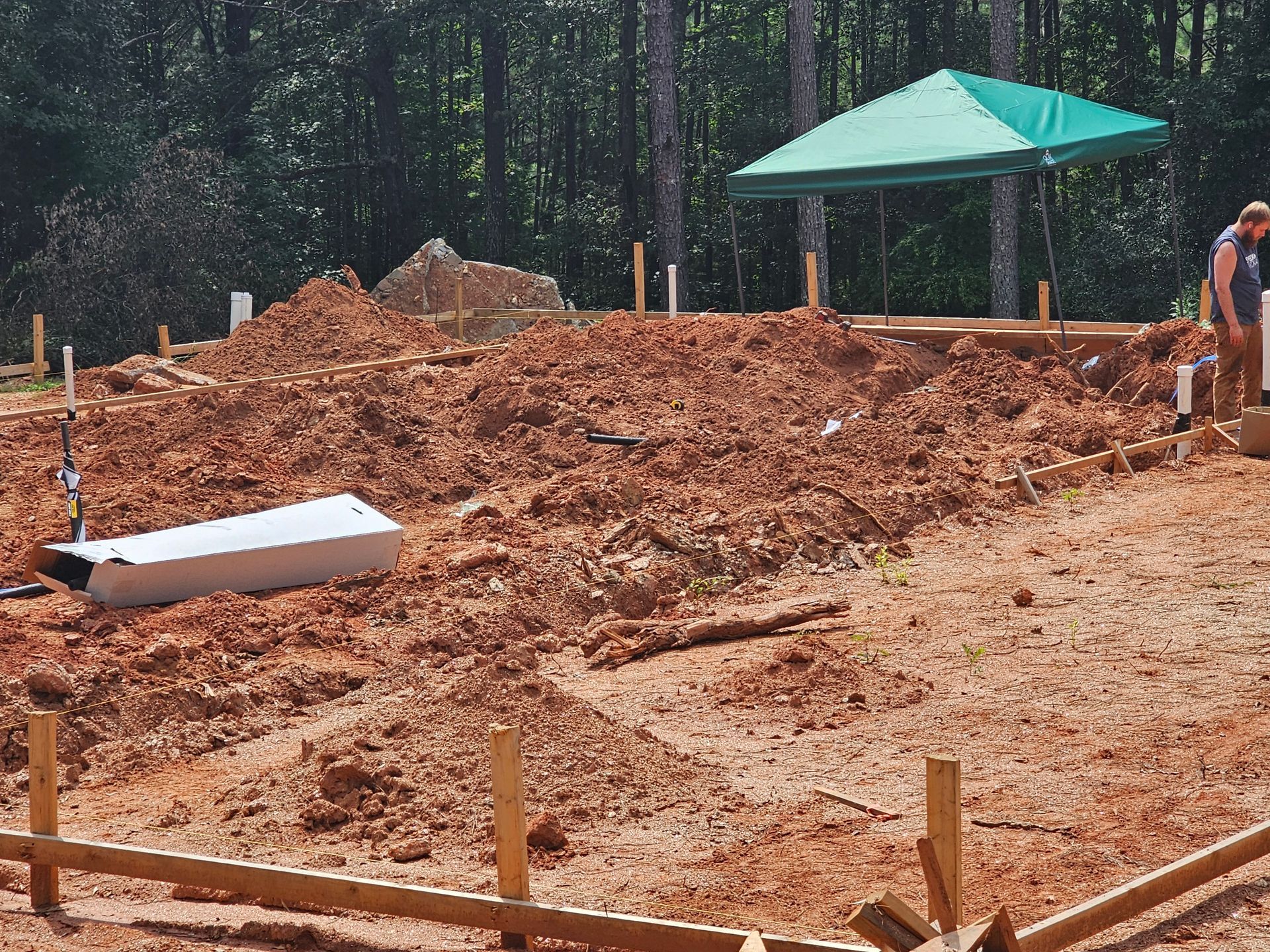 Construction site with a pile of dirt, wooden frames, and a green canopy in a wooded area.