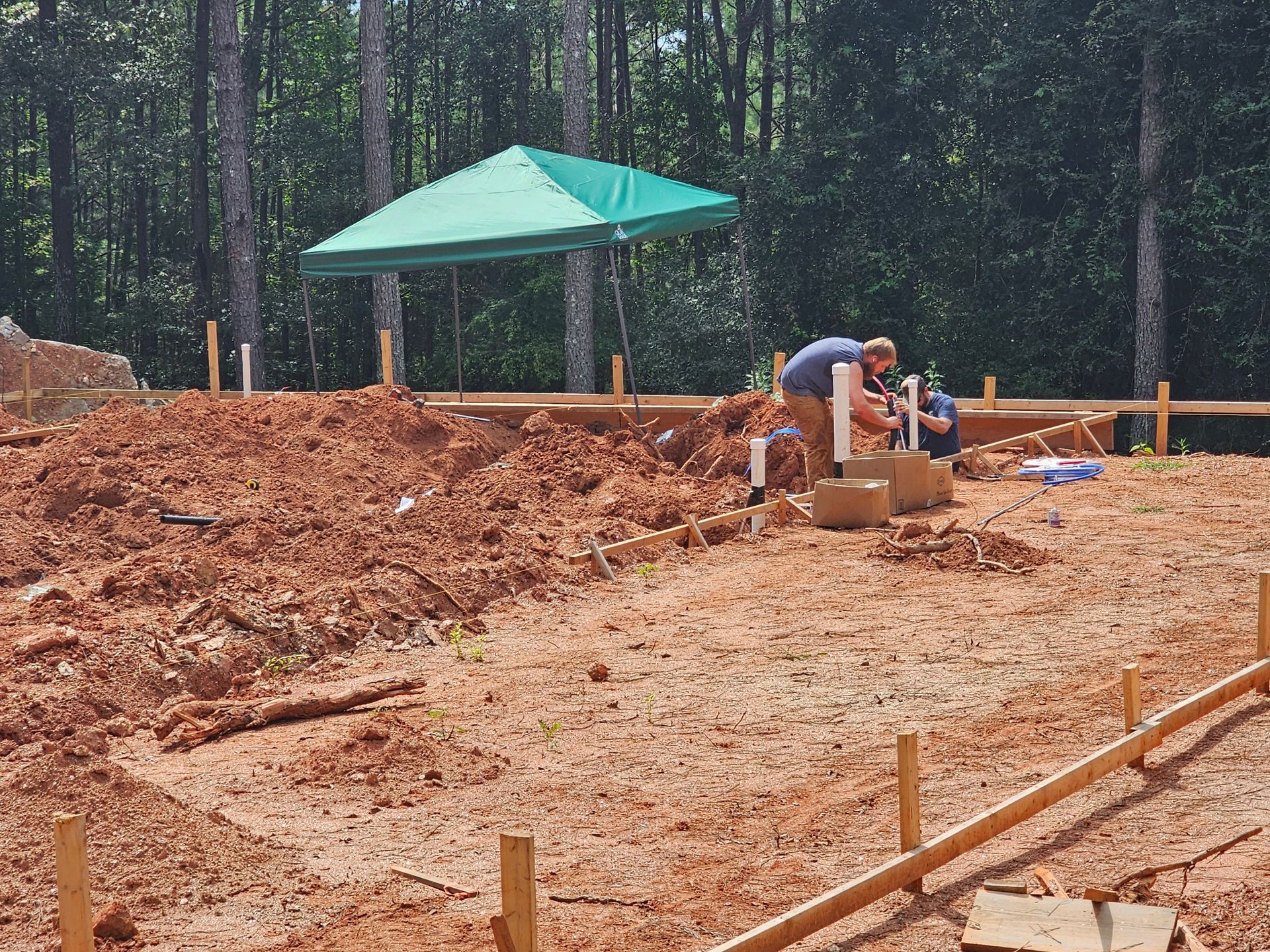 Construction site with workers installing pipes; red dirt, green umbrella, wood frame.