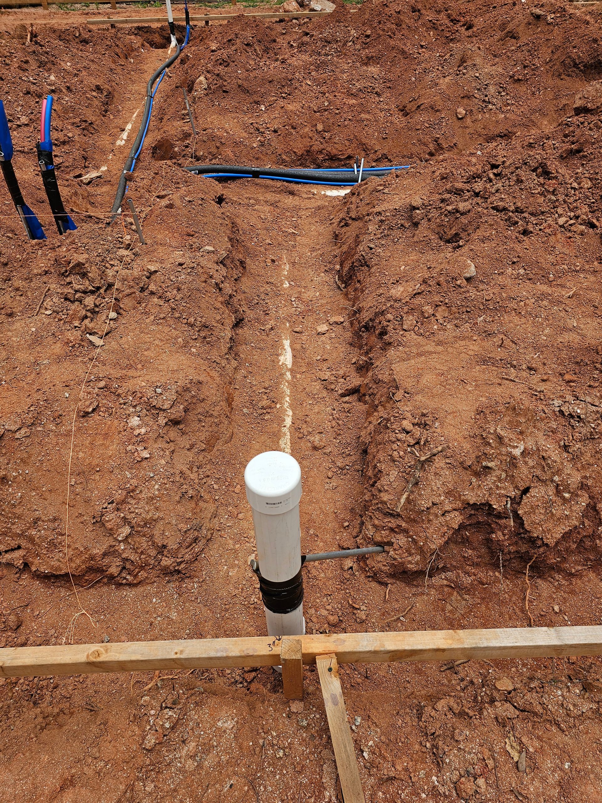 Trench dug in reddish soil with PVC pipe extending upward, blue pipes, and wooden framing.