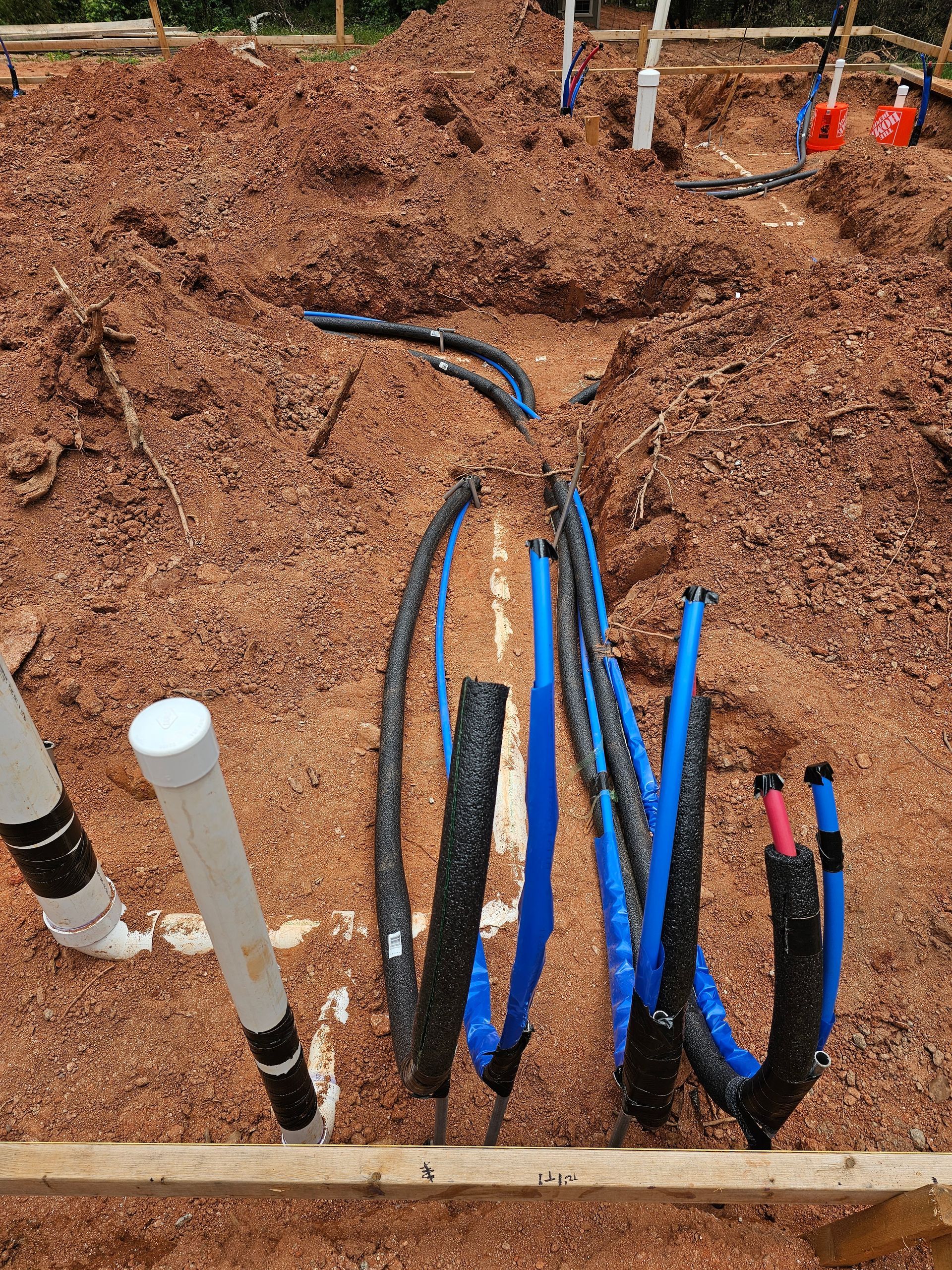 Construction site with blue and black flexible pipes and white PVC pipes in a trench filled with red-brown soil.