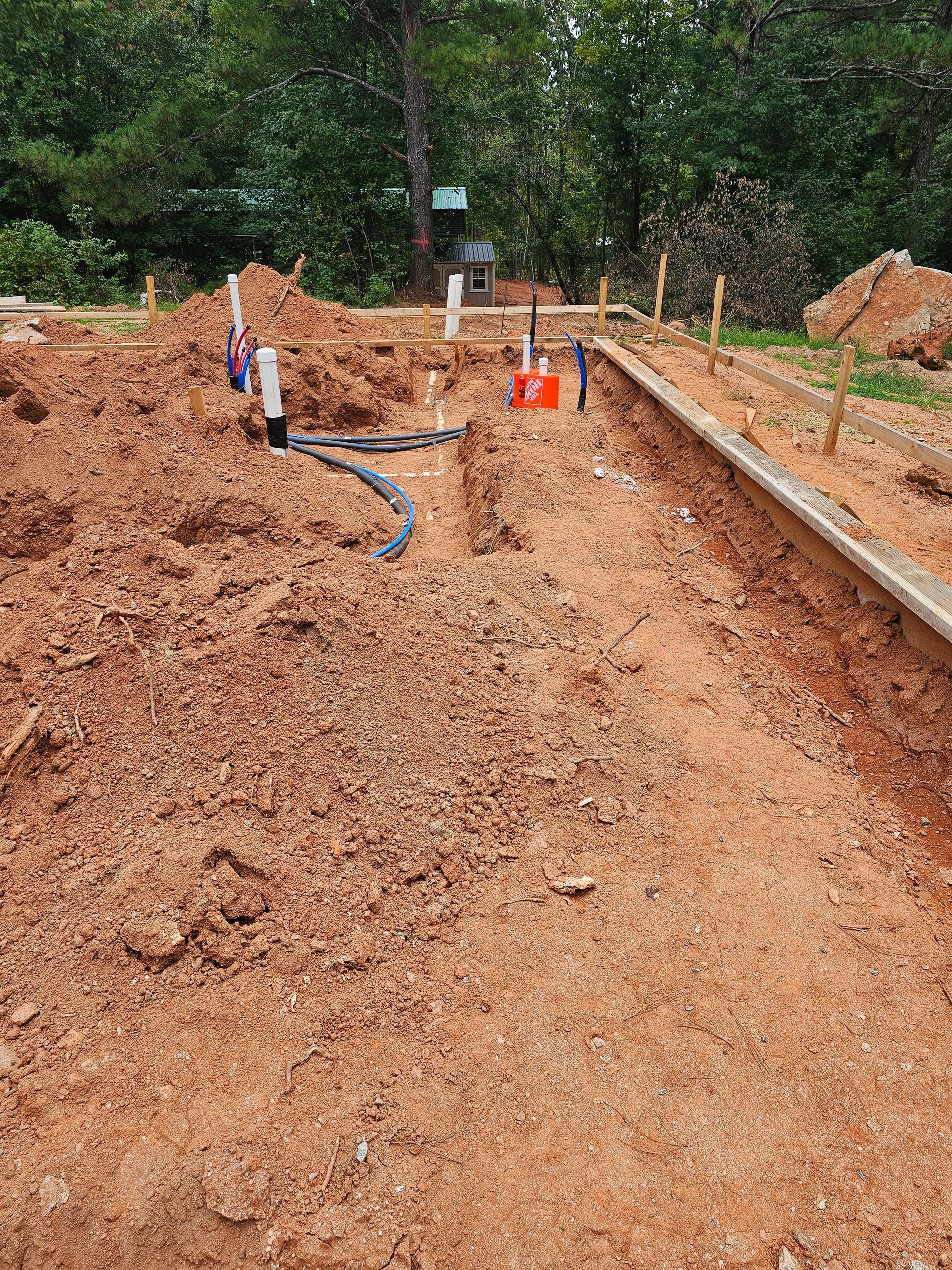 Construction site with exposed plumbing in dirt, wooden border, trees in the background.