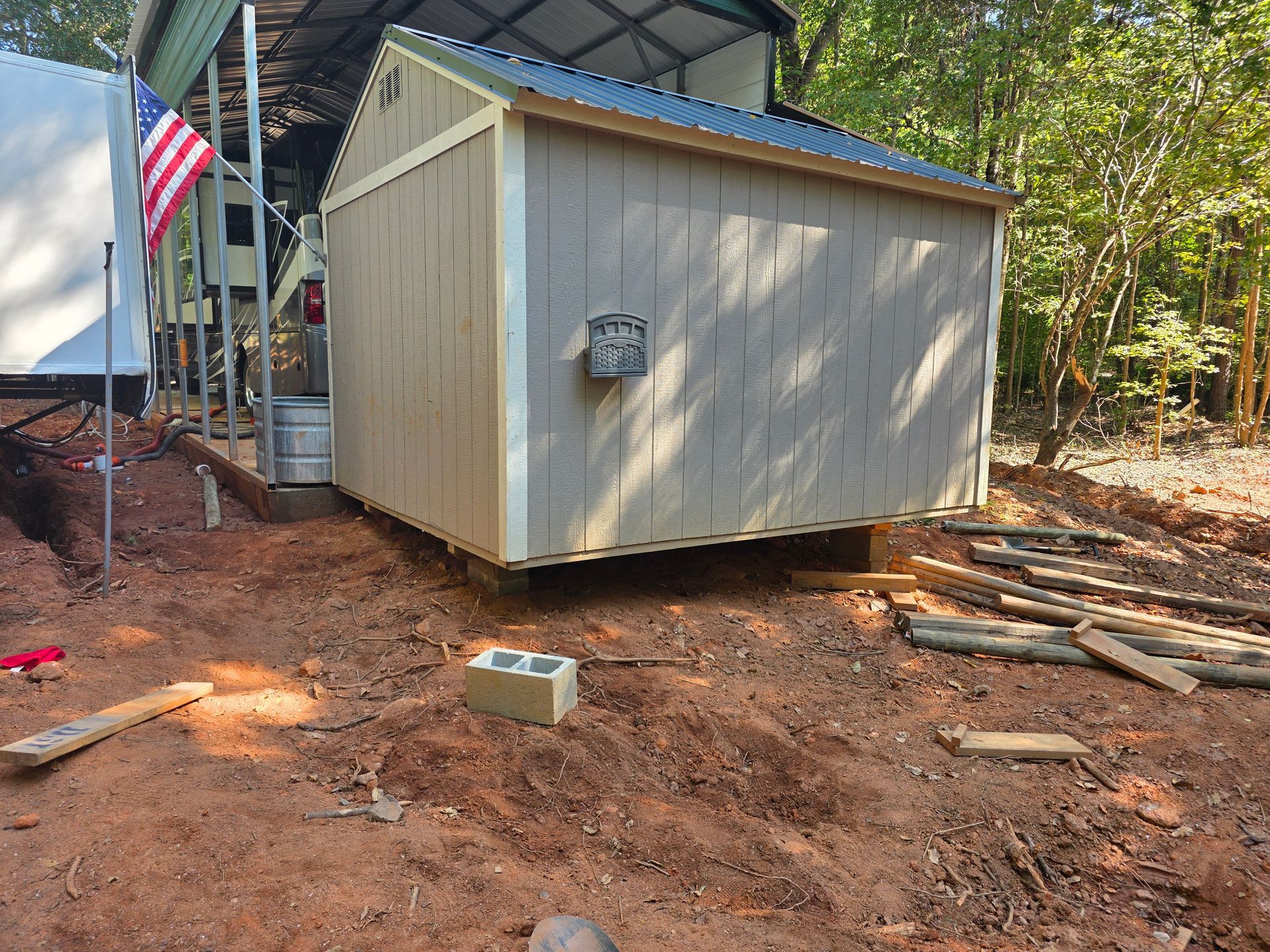 Tan shed on cinder blocks outdoors with an American flag to the left.