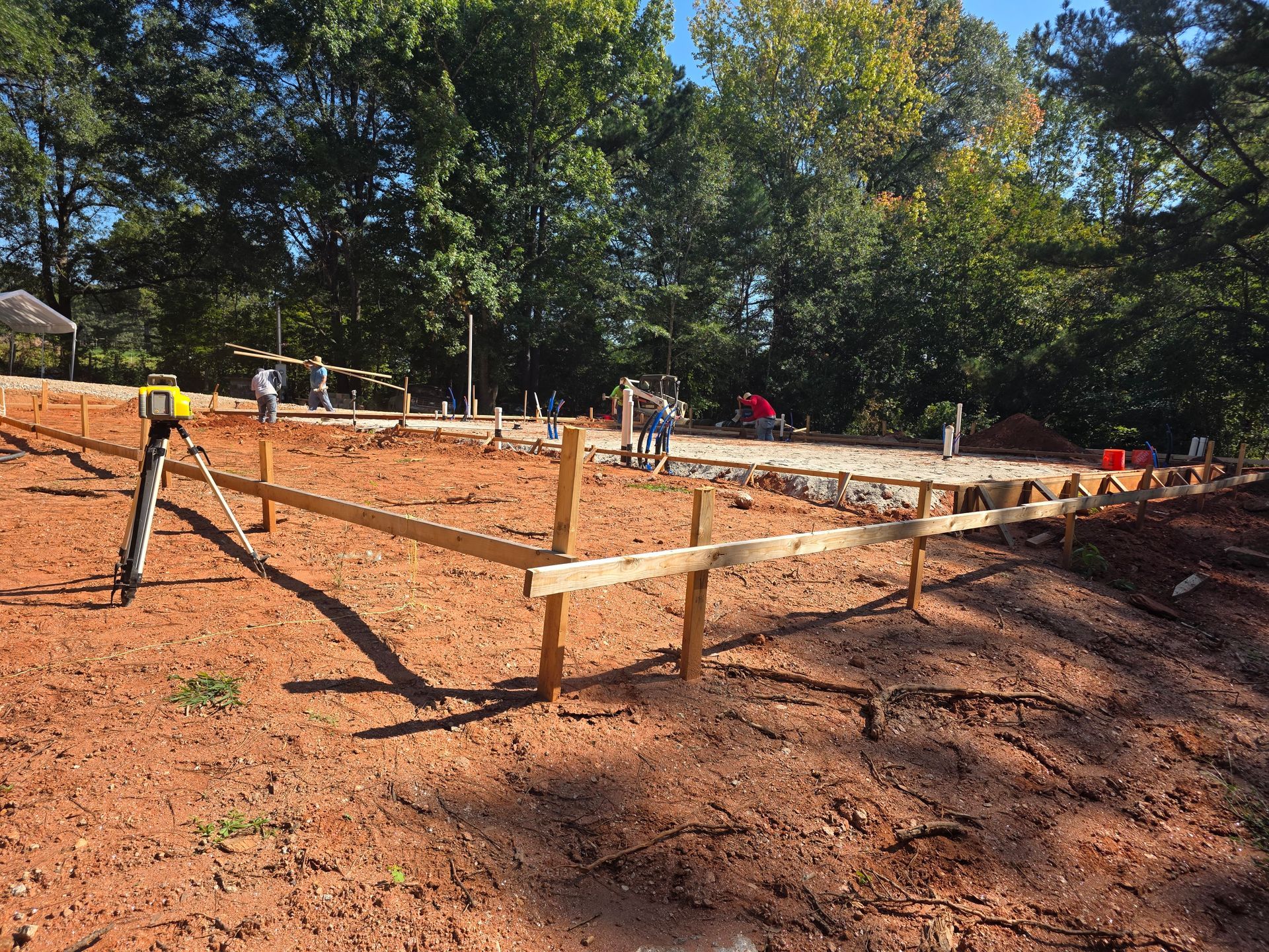 Construction site with wooden forms and workers leveling the ground. Trees in the background.