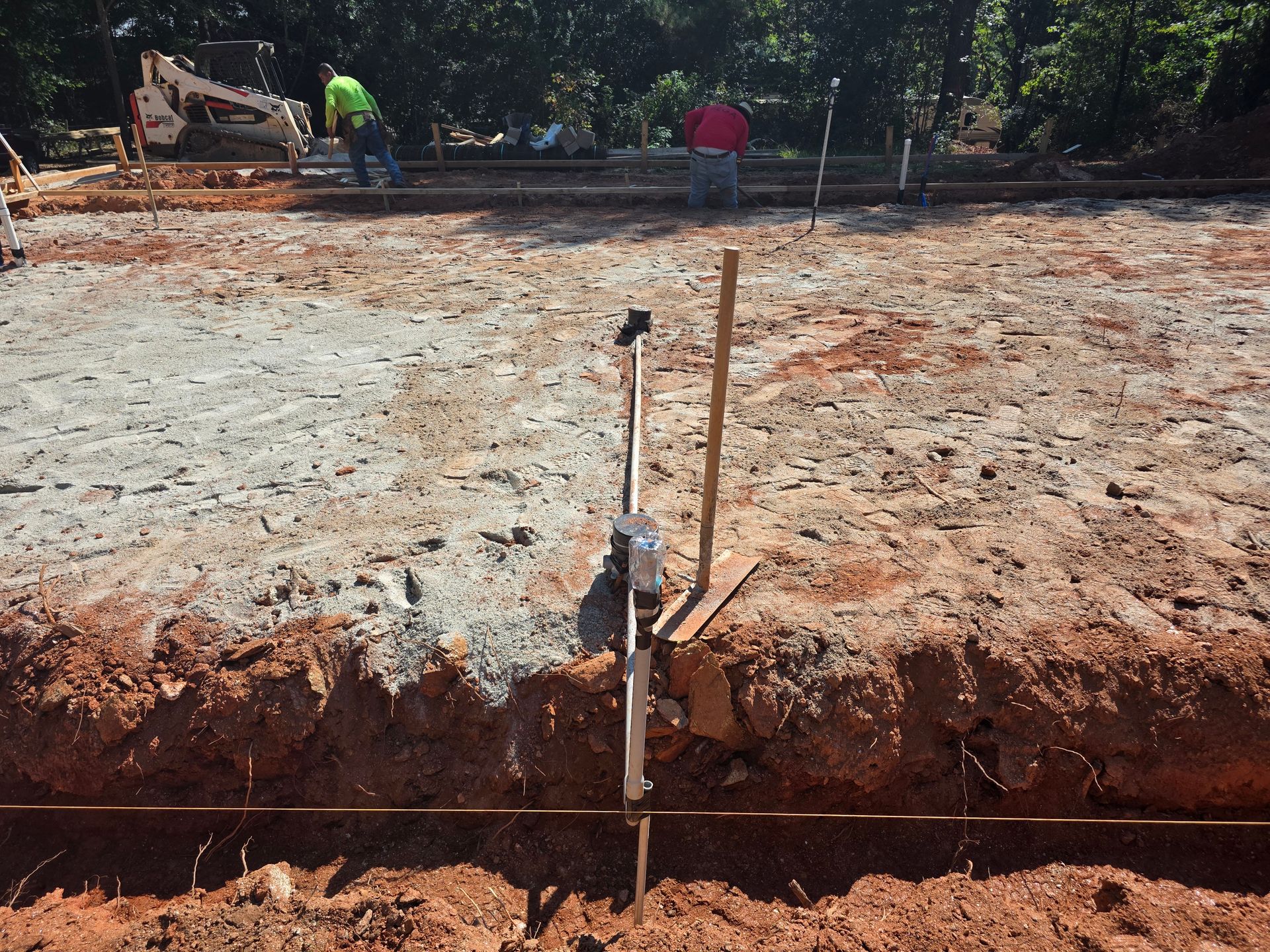 Construction site with workers preparing to pour concrete; leveling tool in foreground, red dirt, and gray ground.
