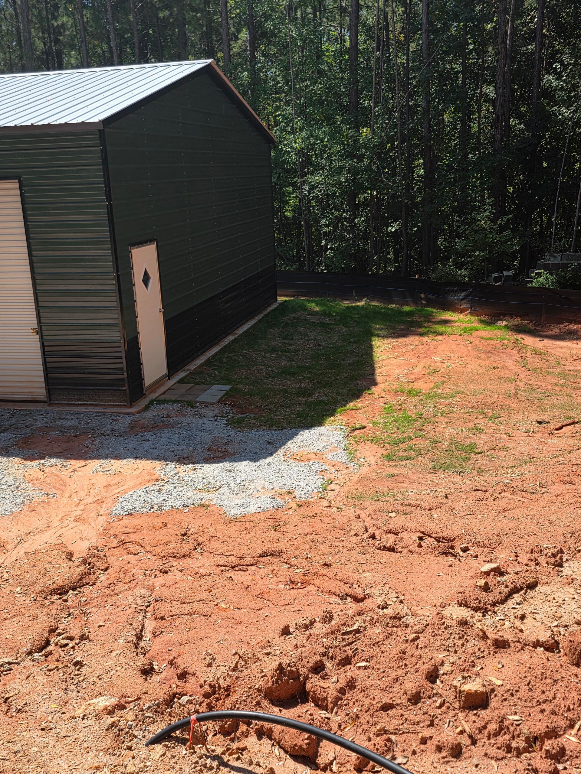 A metal shed with a gravel driveway and red dirt foreground. Sunlight casts a shadow.
