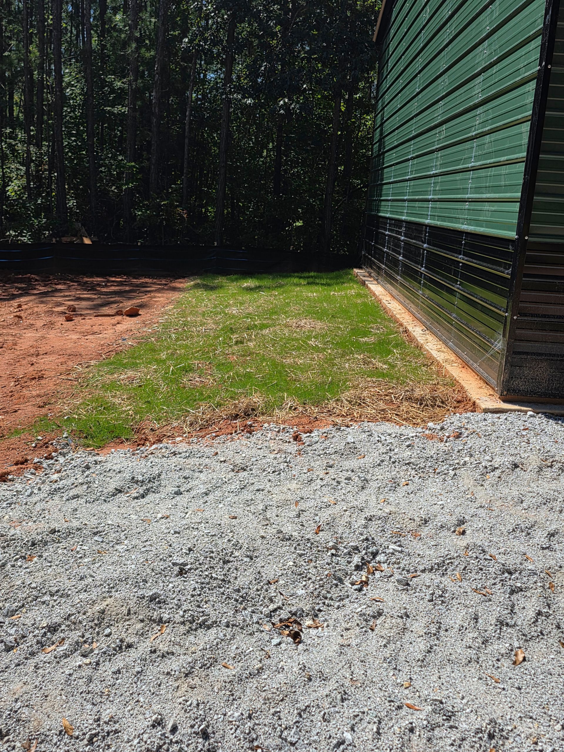 Gravel pathway next to a grassy area, a red-dirt section, and a green building with black siding.