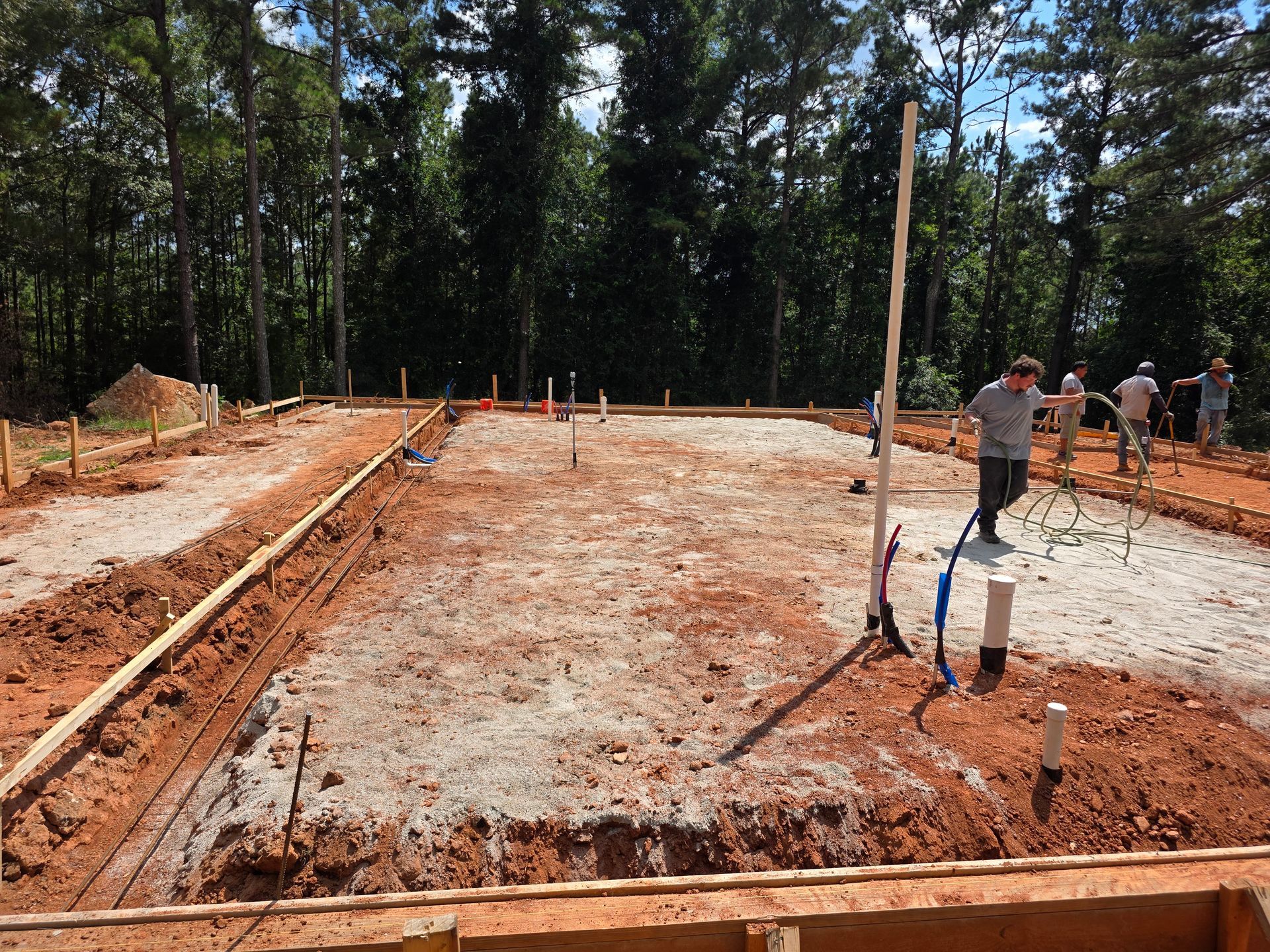 Construction site with wooden frame and trench, workers, plumbing visible, and trees in the background.