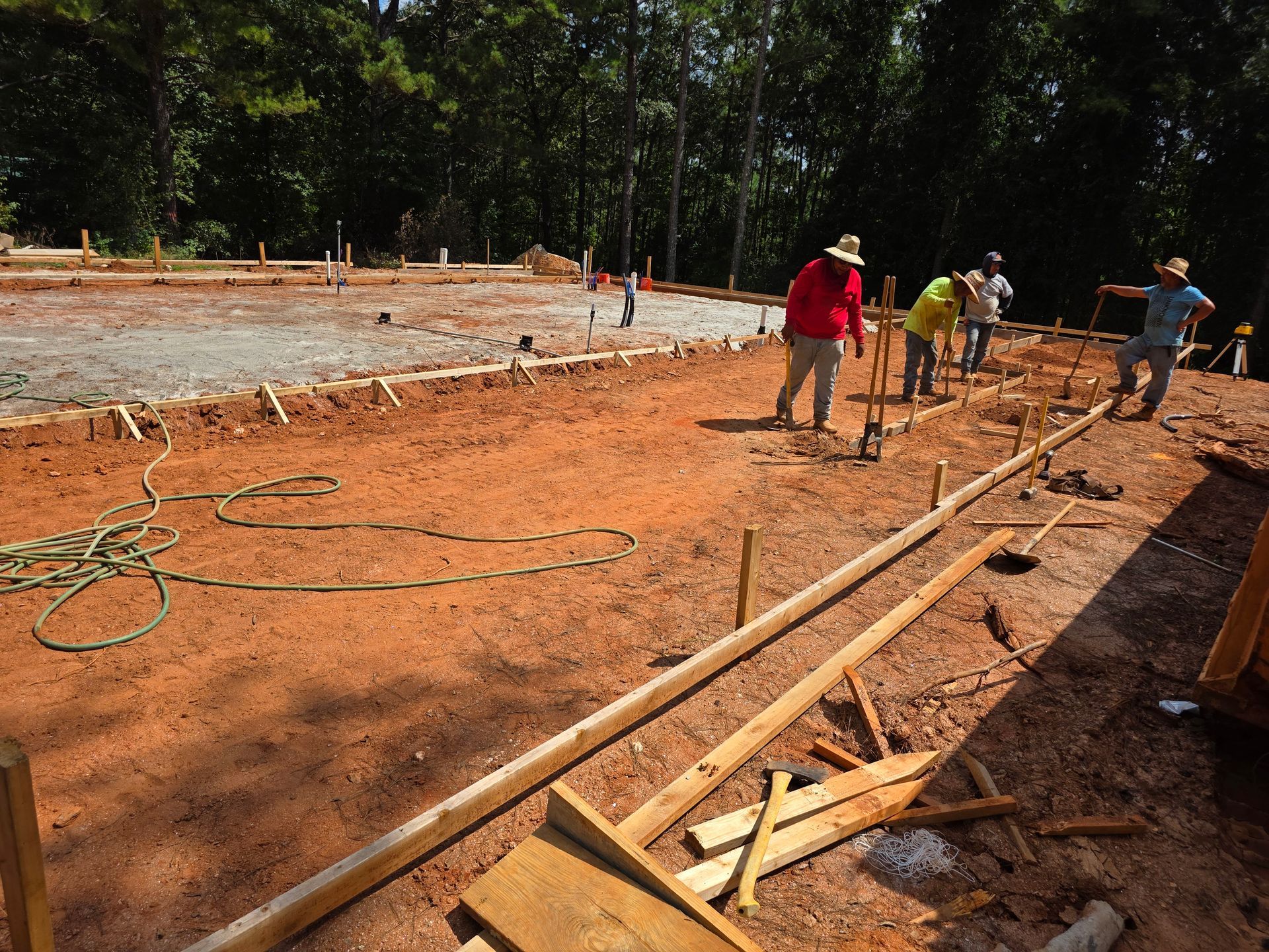 Construction workers leveling ground at a job site with wood forms, surrounded by trees.