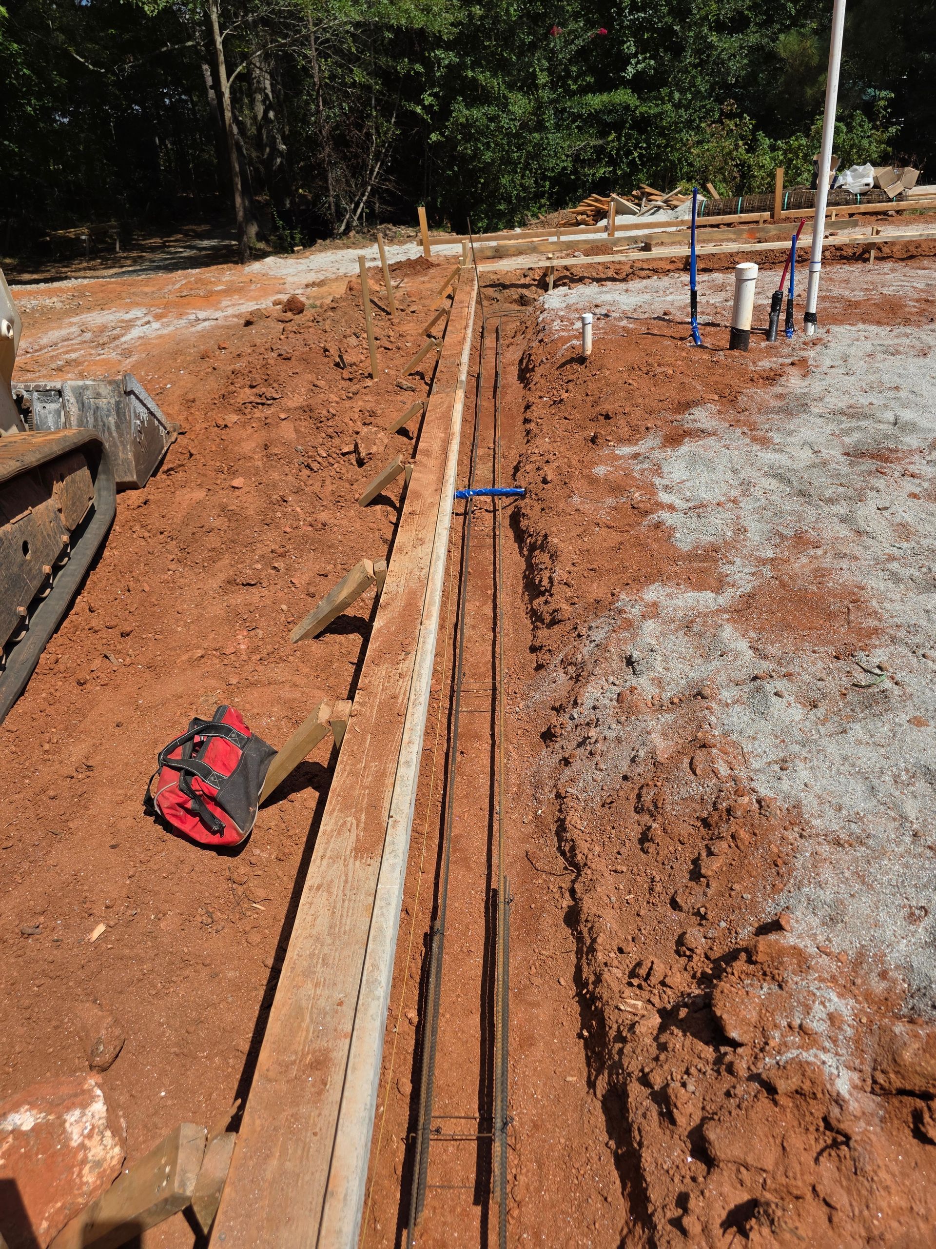 Construction site with concrete forms, rebar, and plumbing pipes set in reddish soil.