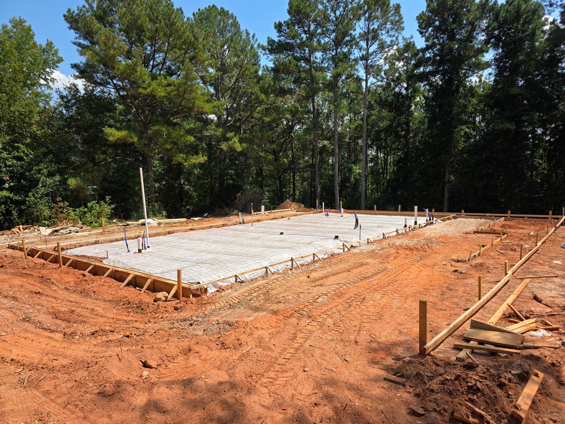 Construction site with concrete foundation and plumbing, surrounded by dirt and trees.