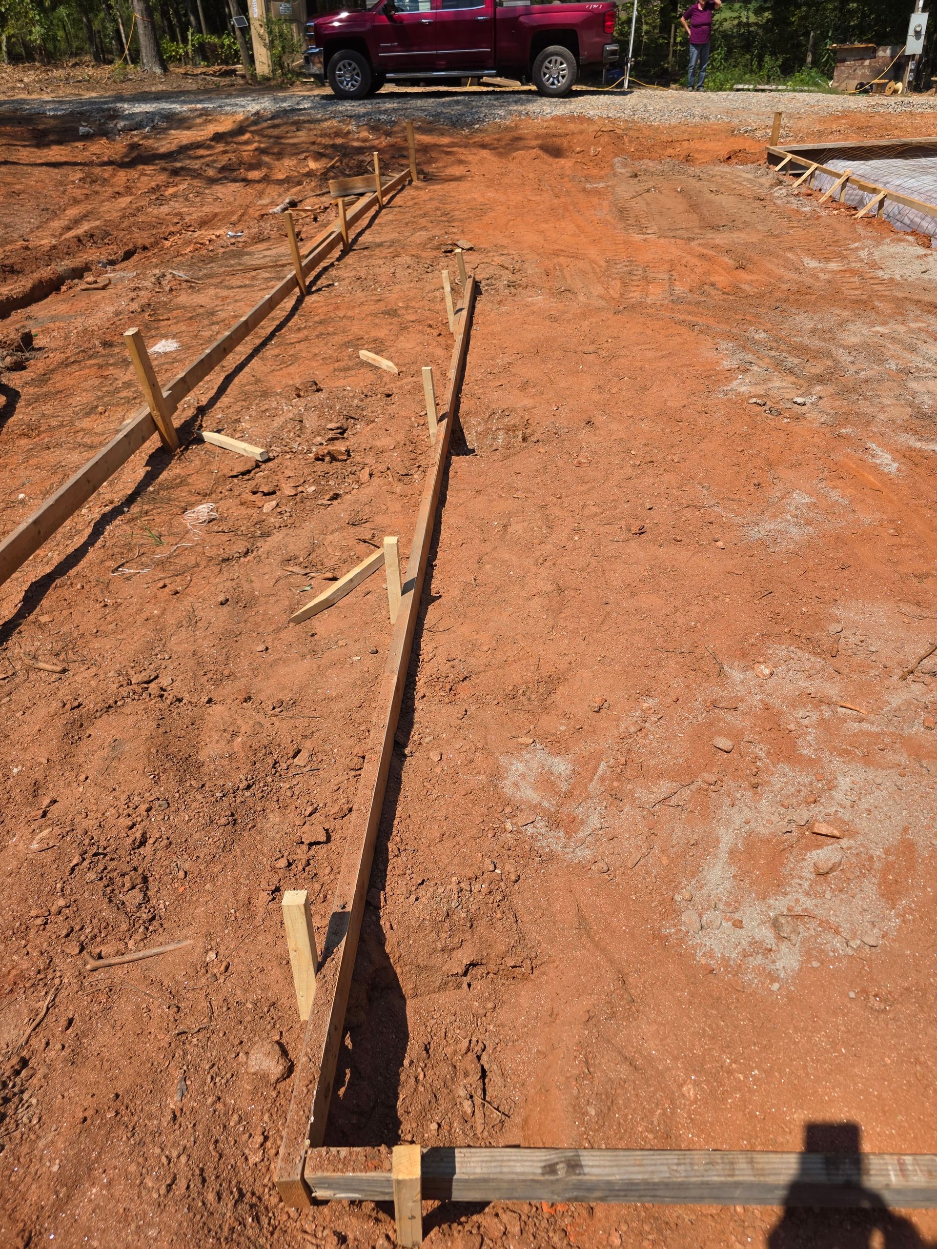 Rows of wooden stakes guide construction on red dirt ground, truck in the distance.