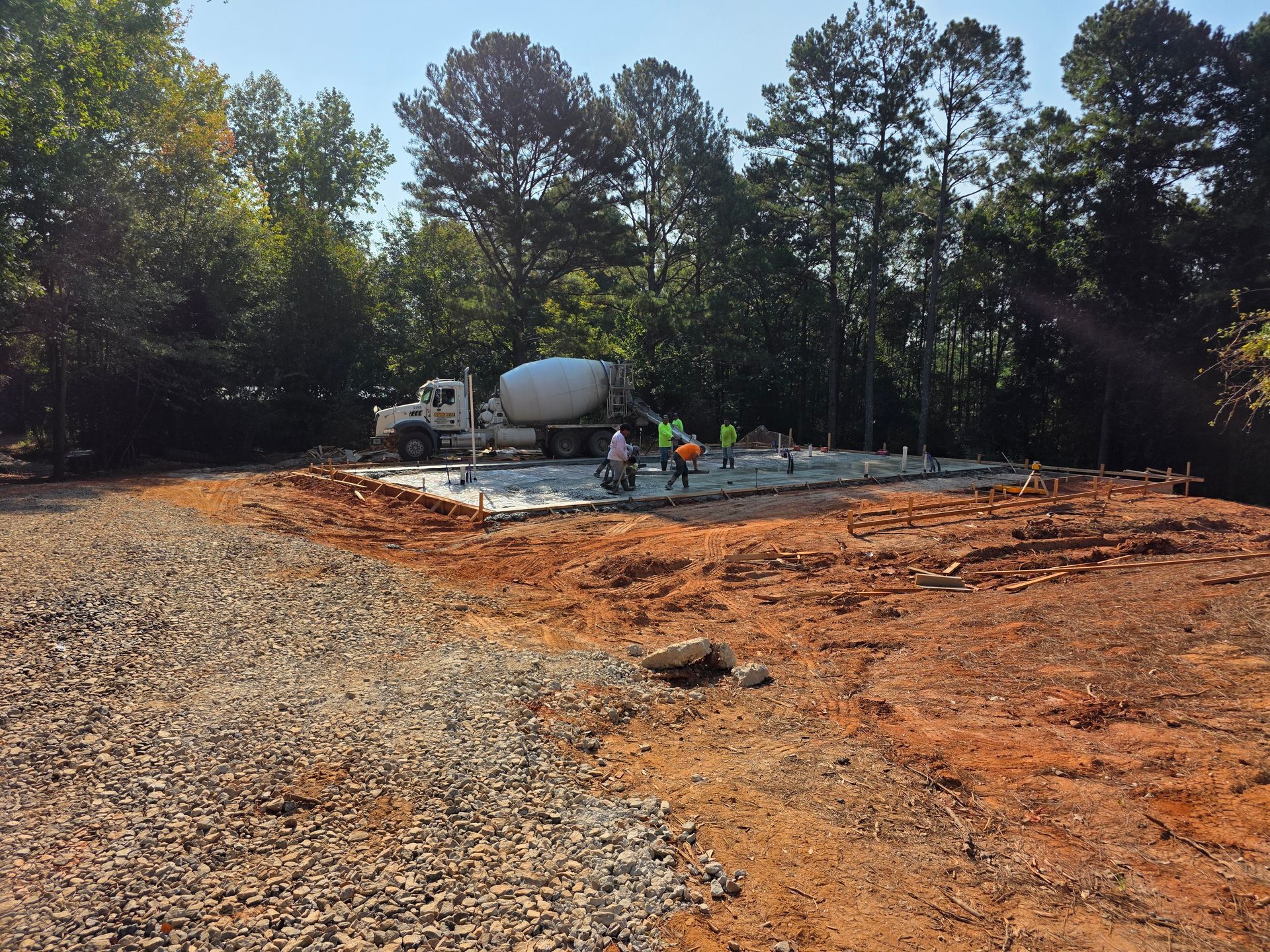 Concrete pouring for foundation, with truck and workers on a leveled dirt lot. Trees in the background.
