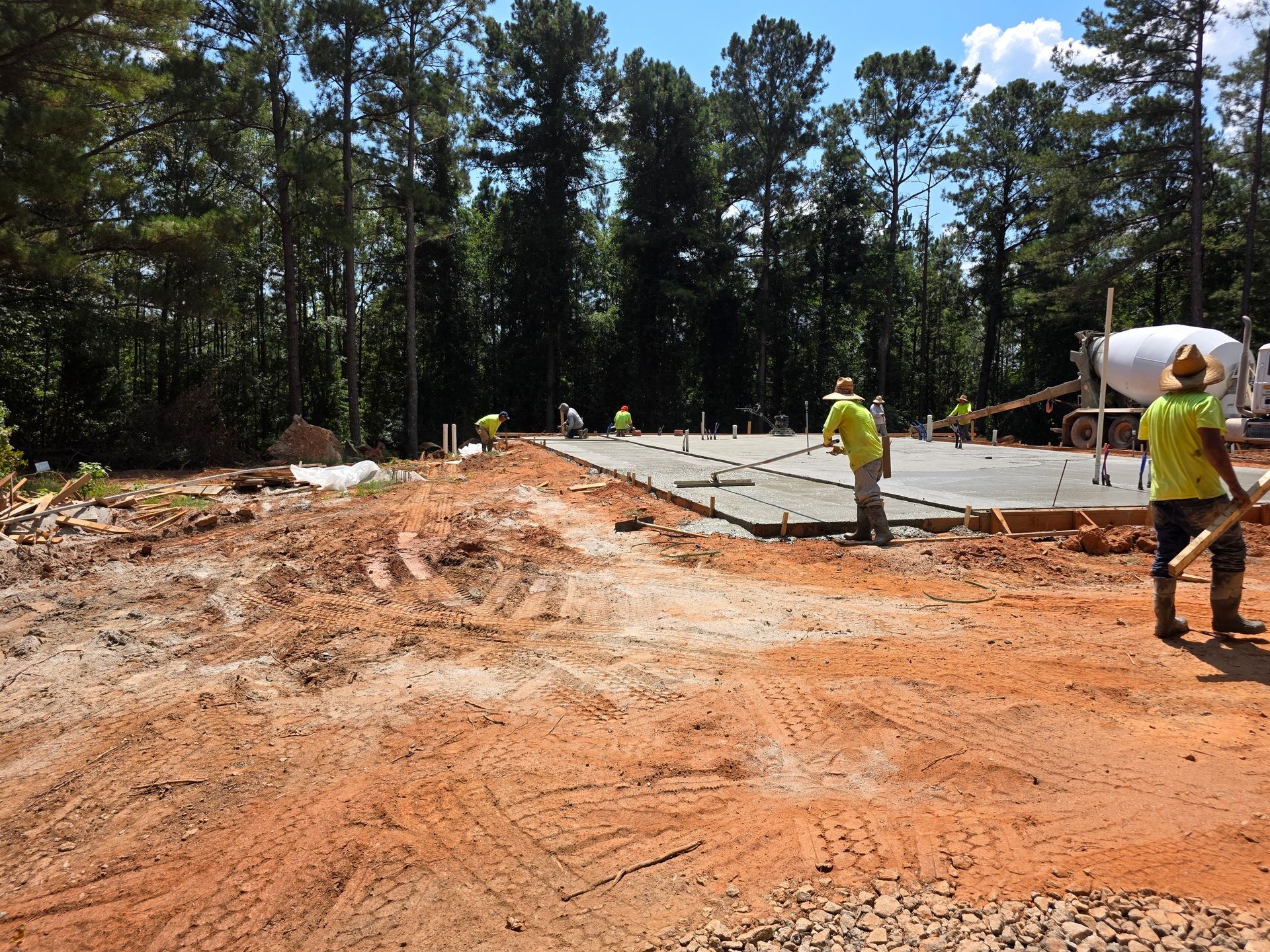 Construction workers pour concrete slab for a building foundation in a wooded area. Workers wear yellow shirts and hard hats.