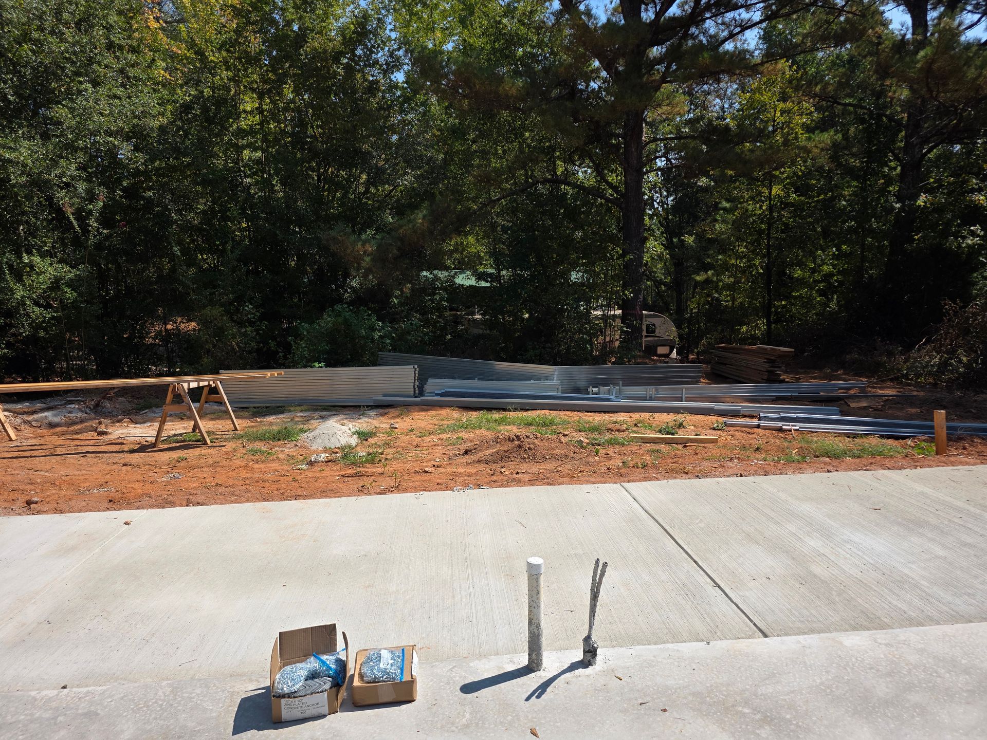 Construction site with lumber and supplies on a dirt patch, in front of trees, and a concrete driveway in the foreground.