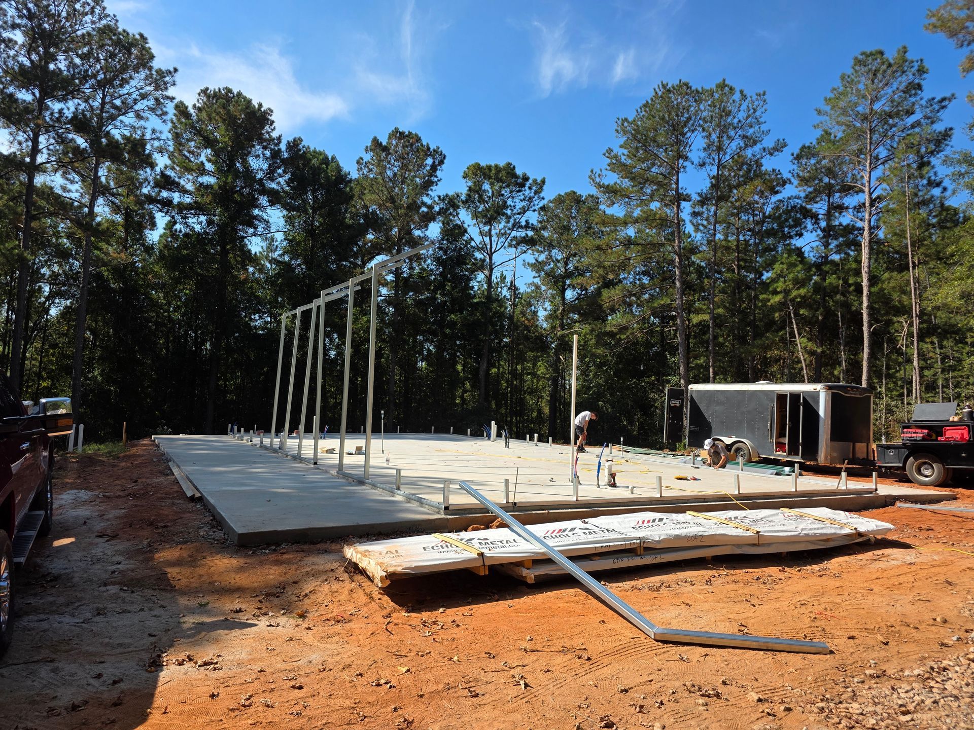 Construction site: concrete foundation with steel framework, trees, and blue sky.