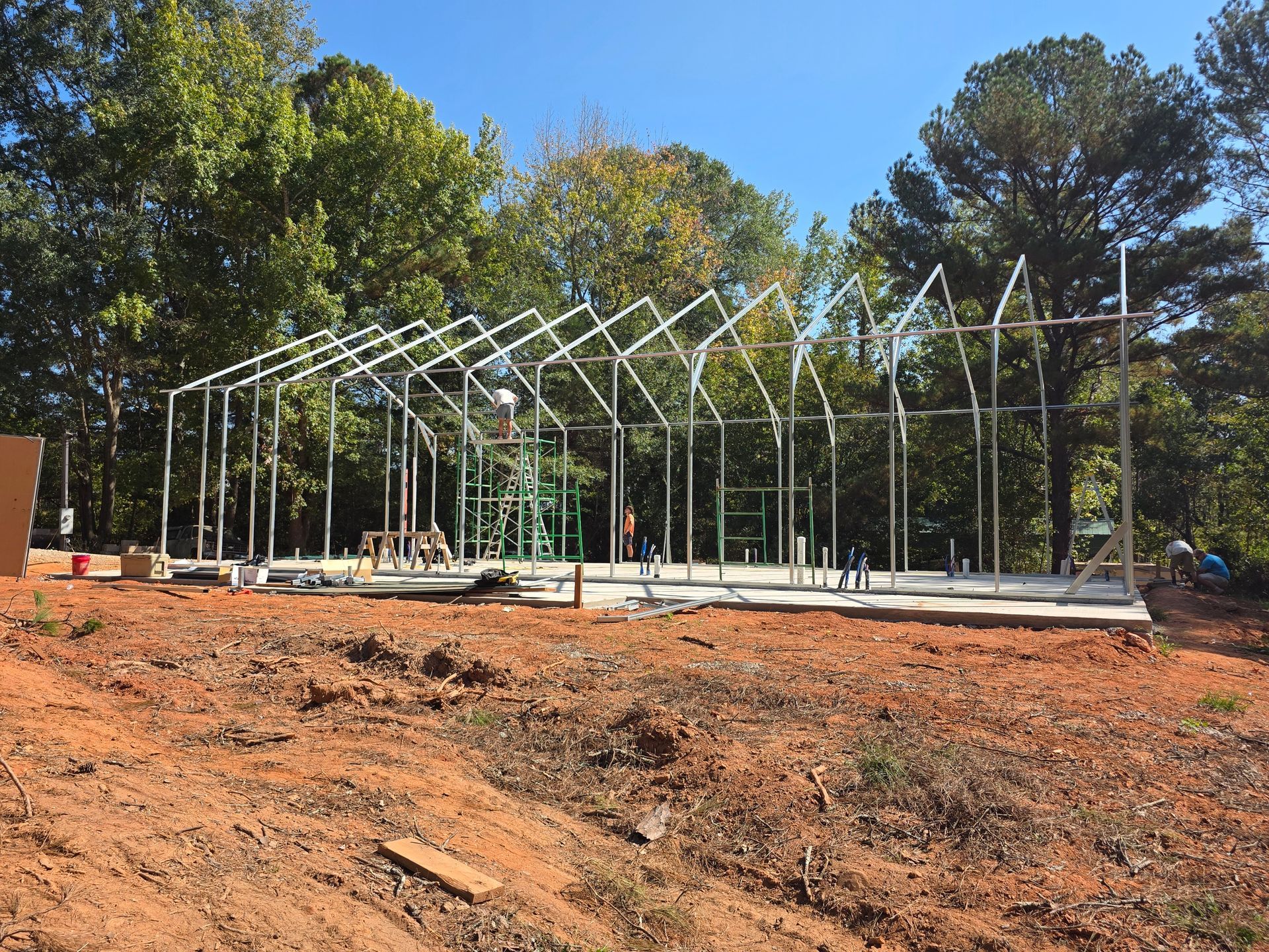 Construction site with a metal frame building in progress, set on a concrete slab in a wooded area.