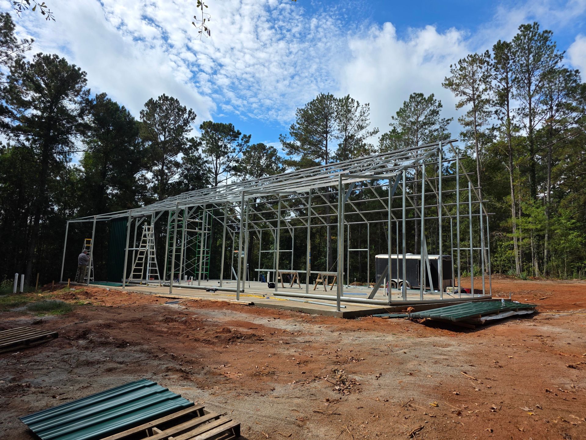 Steel frame of a building under construction on a dirt lot, surrounded by trees.