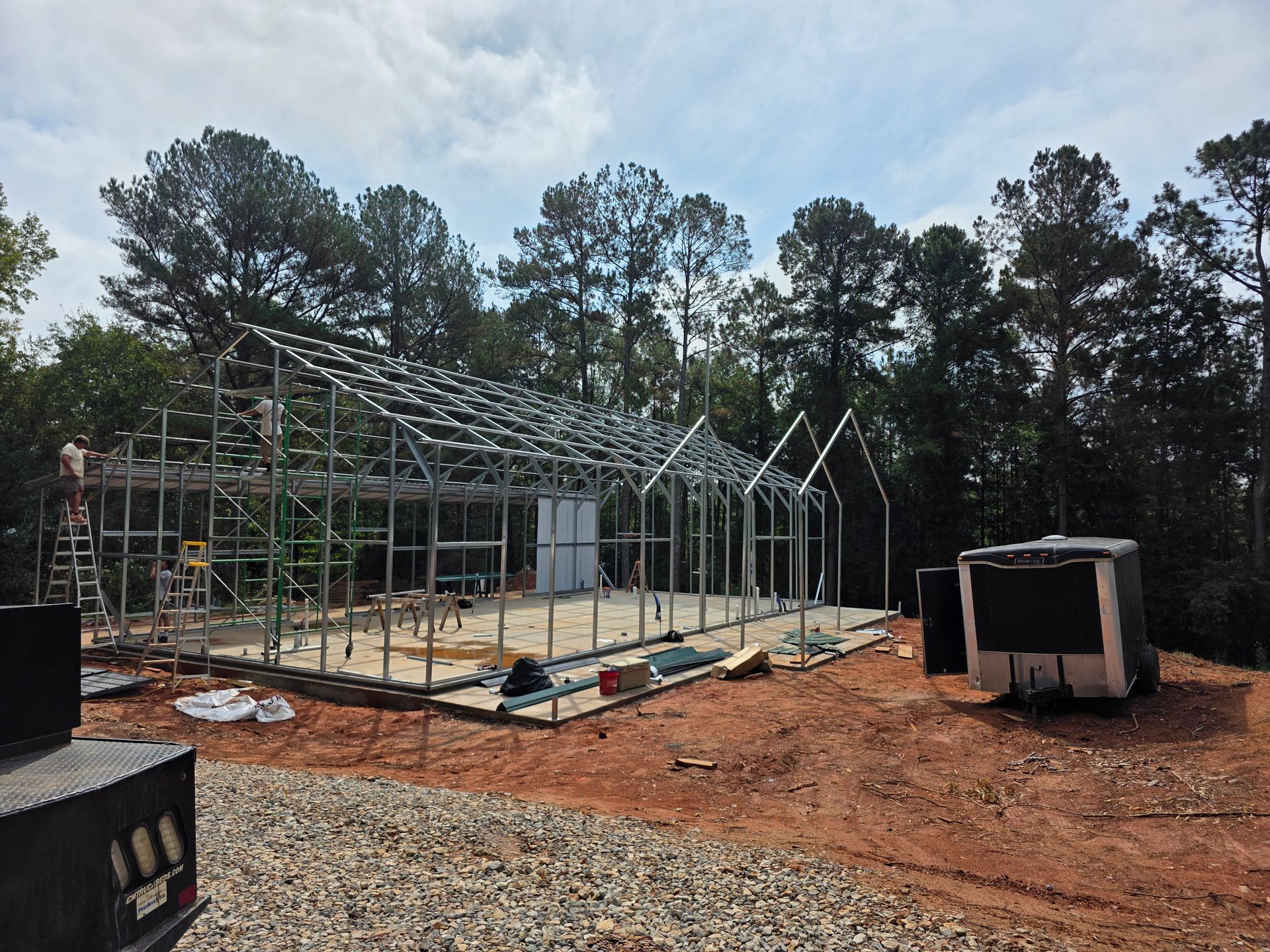 Steel frame of a building under construction on a dirt lot, surrounded by trees.