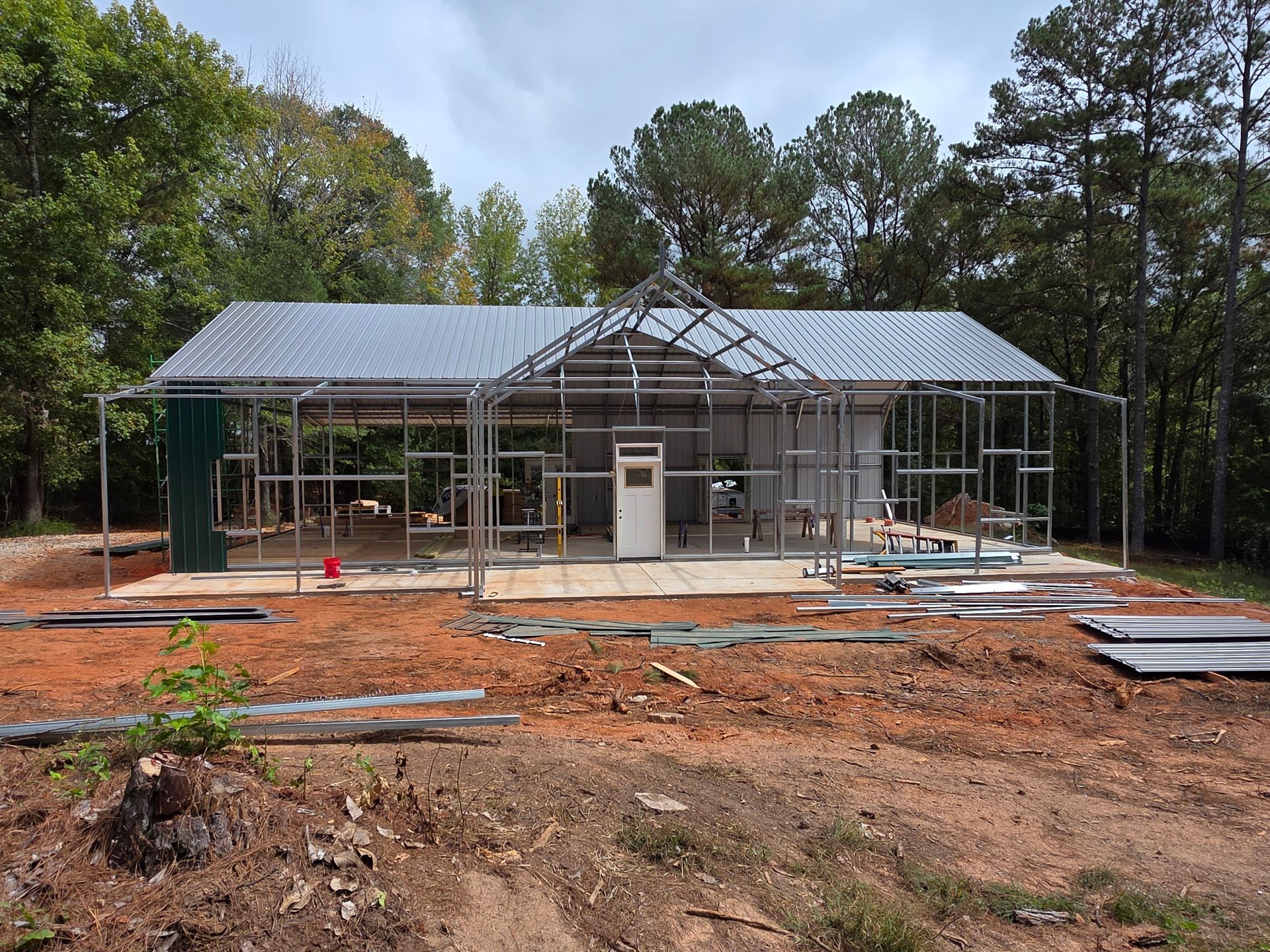 Metal frame building under construction, partially enclosed with green and silver panels, set in a wooded area.