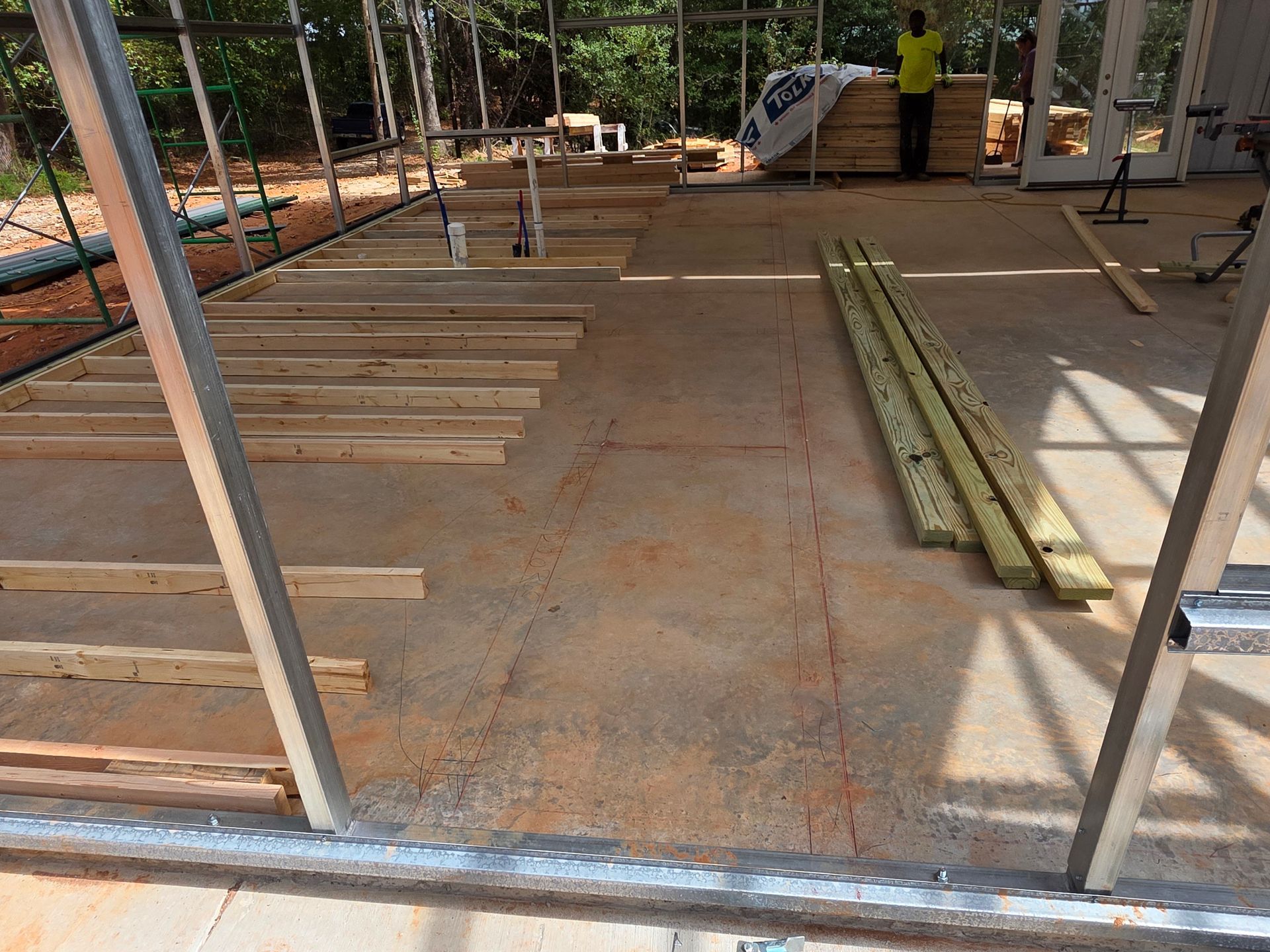 Construction site interior with lumber and metal framing on concrete floor. A worker stands in the background.