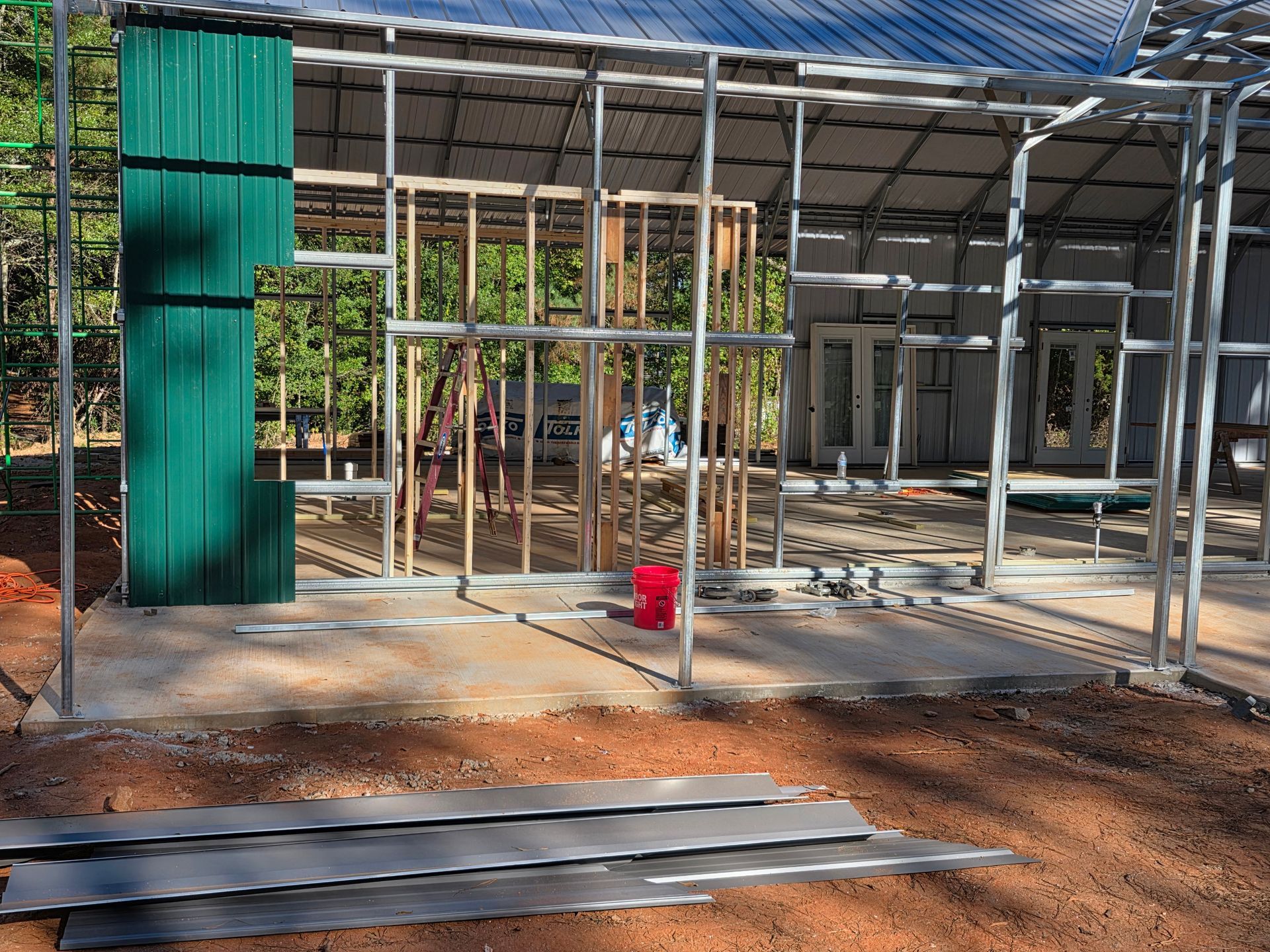 Construction site with metal framing and green siding, red bucket, and metal sheets.