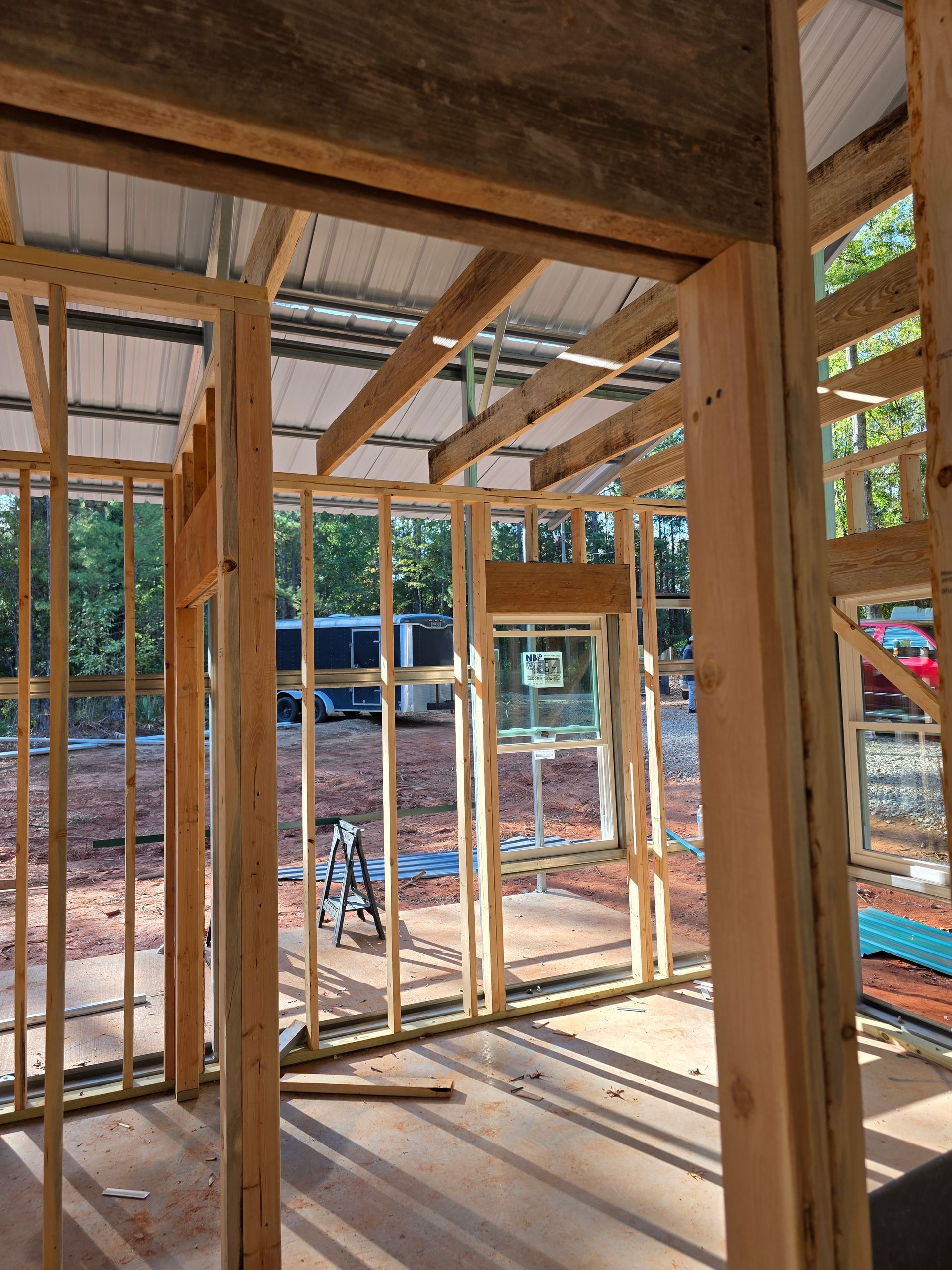 Interior view of a building under construction, framed with wooden studs and rafters, with a metal roof.
