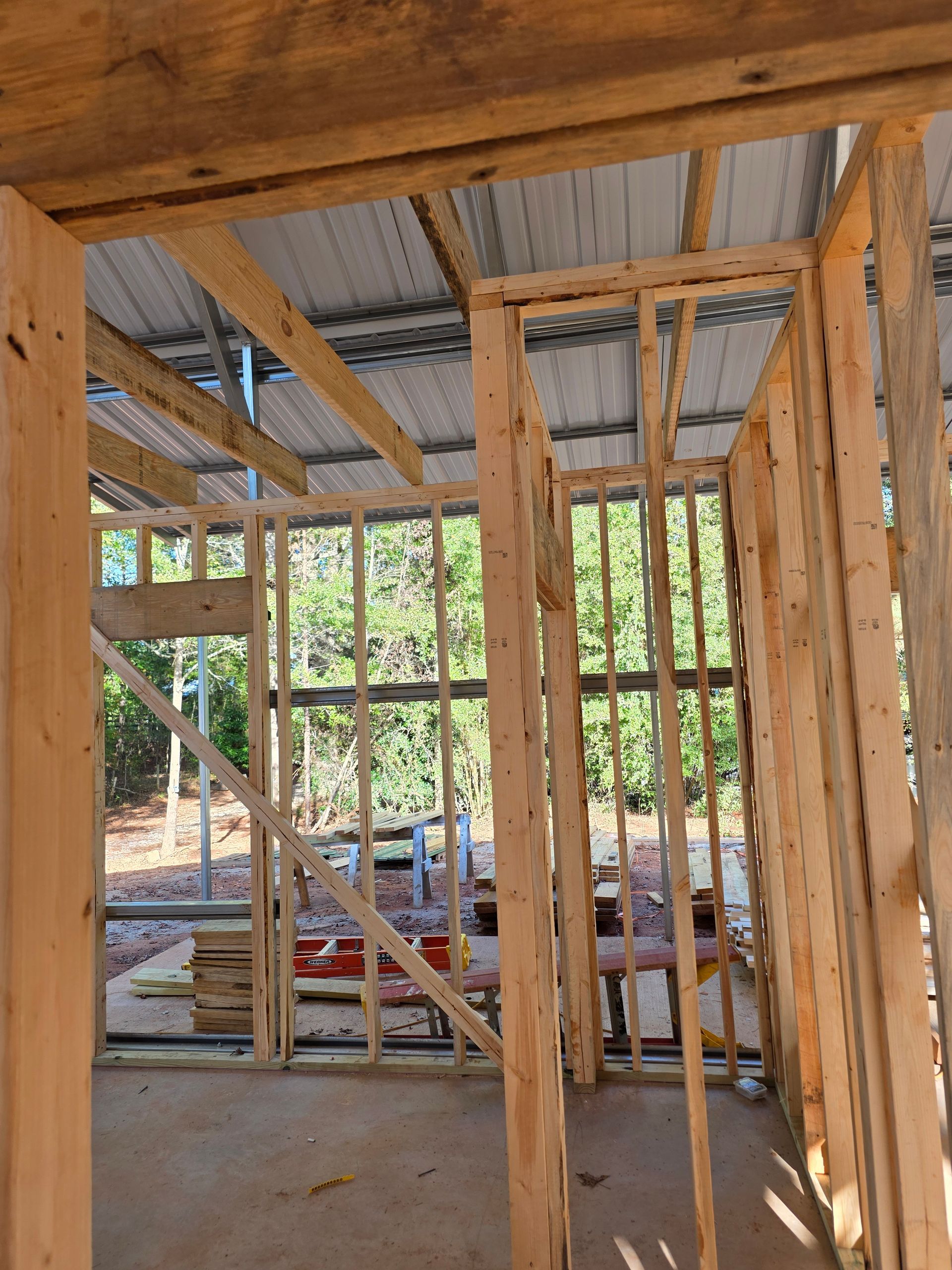 Wooden frame construction of a building, with exposed studs and rafters; metal roof visible, outdoors.