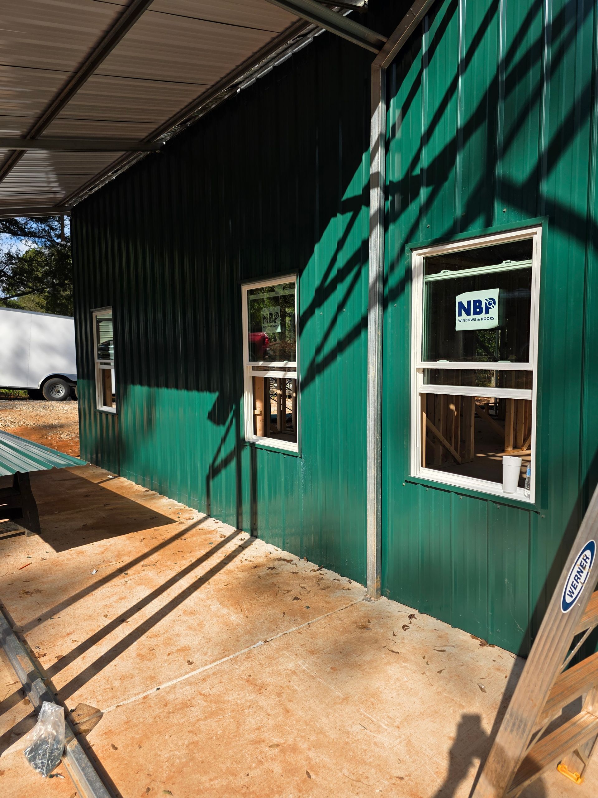 Green metal-sided building with three windows, under construction, with a white trailer in the background.